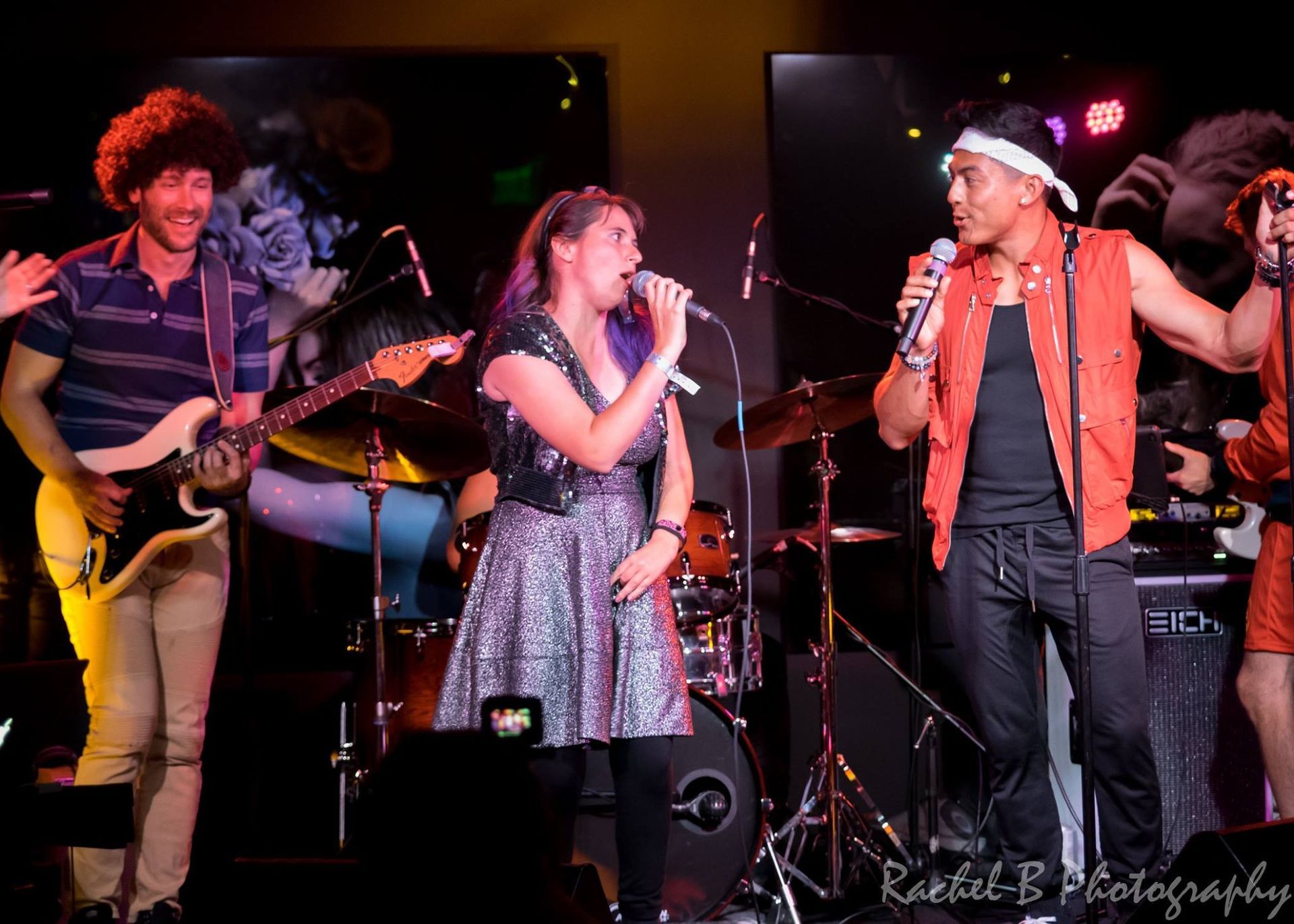 A band performs on stage with two singers at microphones and a guitarist playing a white electric guitar.