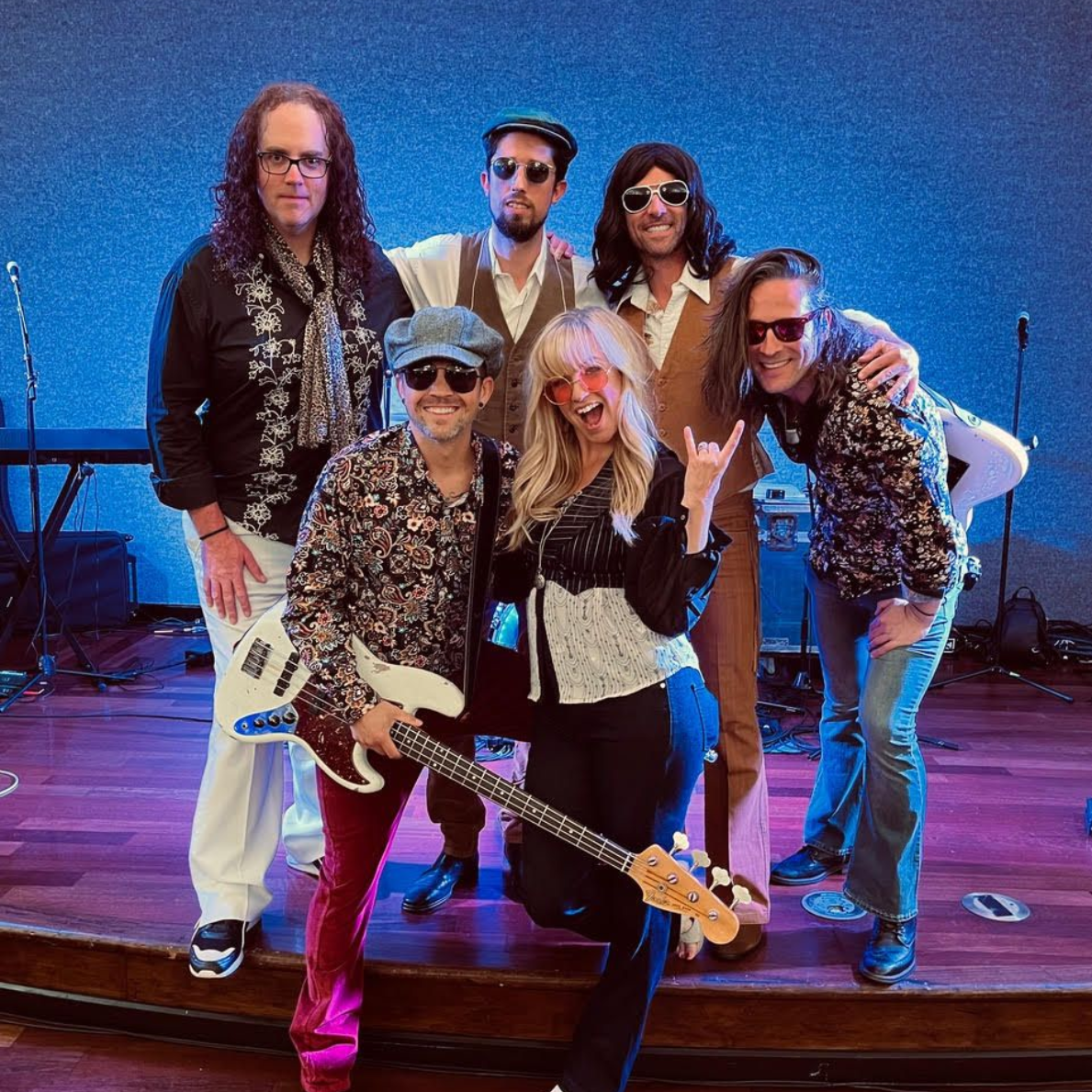 A six-piece band poses on a wooden stage with blue lighting; one member holds a bass guitar while others wear sunglasses.