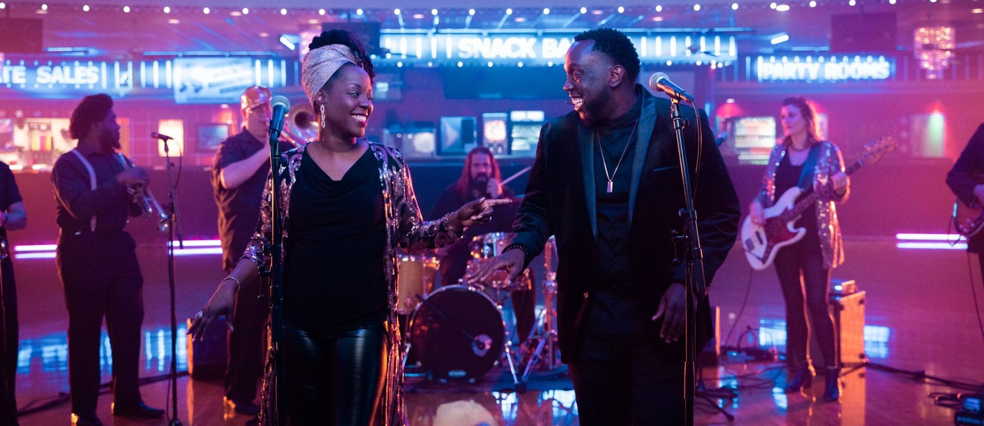 A smiling duo sings into microphones on a stage, backed by a band playing instruments in a dimly lit, red-hued venue.