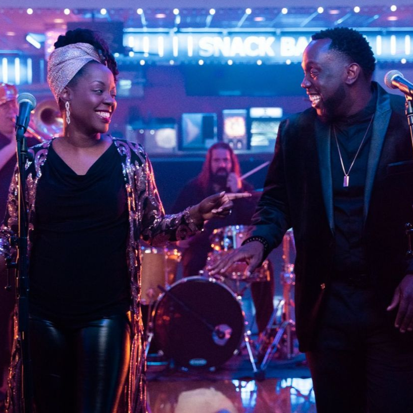 A smiling duo sings into microphones on a stage, with a drummer in the background and a