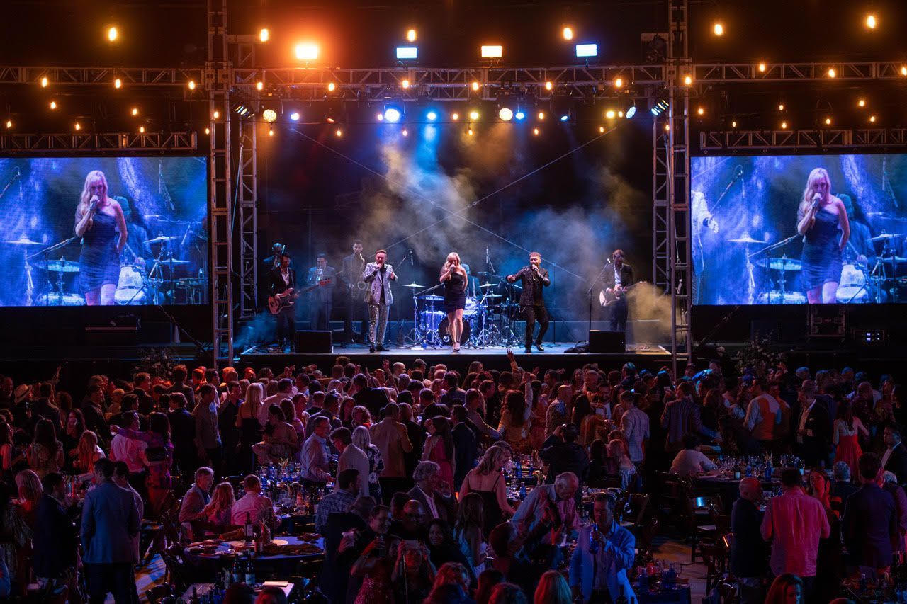A live musical band performs on a concert stage with large side screens, before a dense crowd at an outdoor night event.