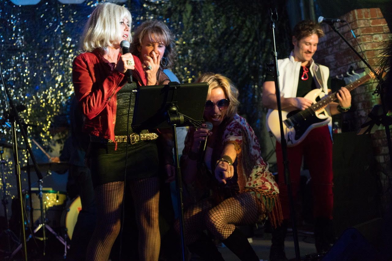 A band performs on stage with two singers at microphones and a guitarist playing a white electric guitar.