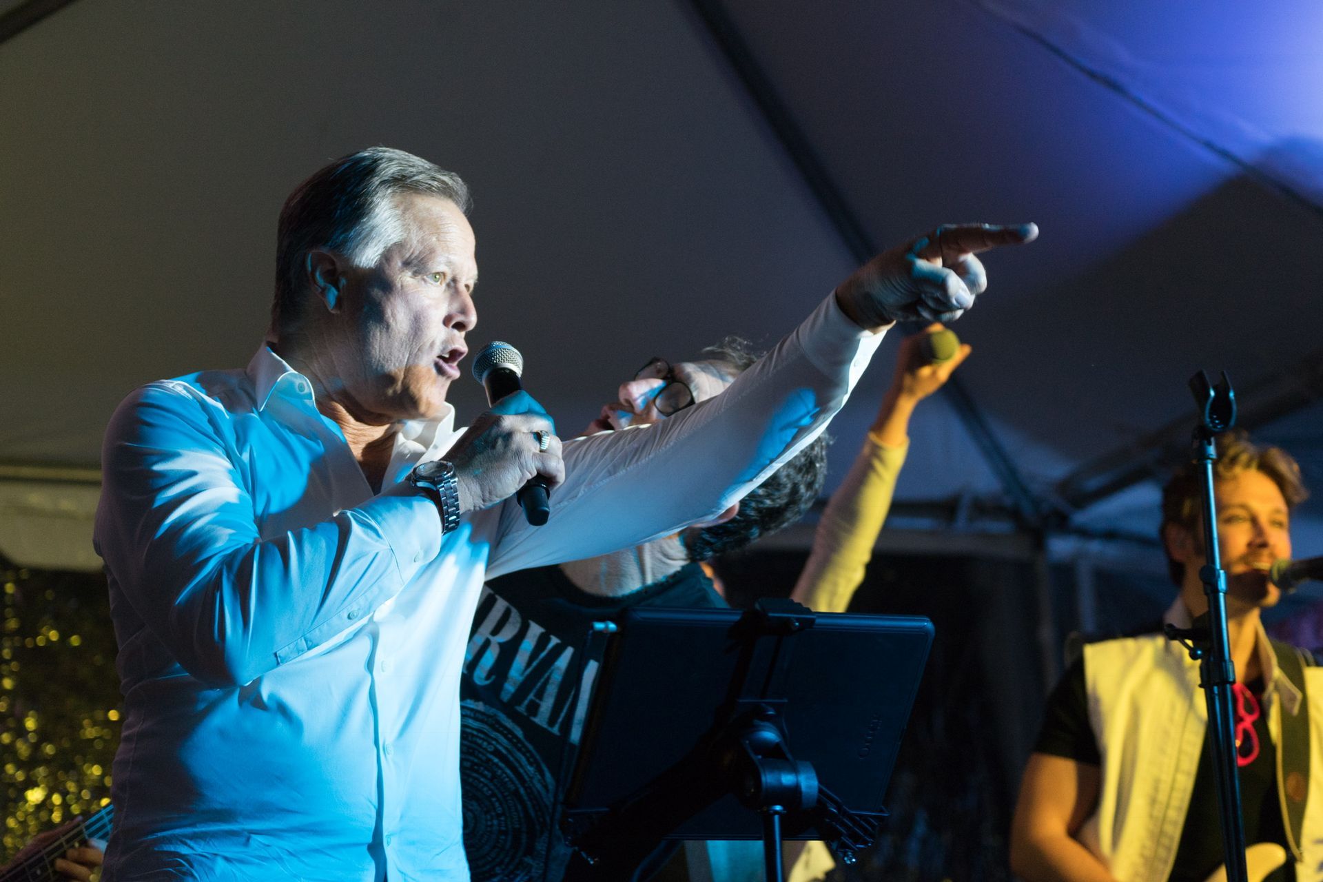 A singer in a white shirt points while performing on stage with a band under a tent at night.