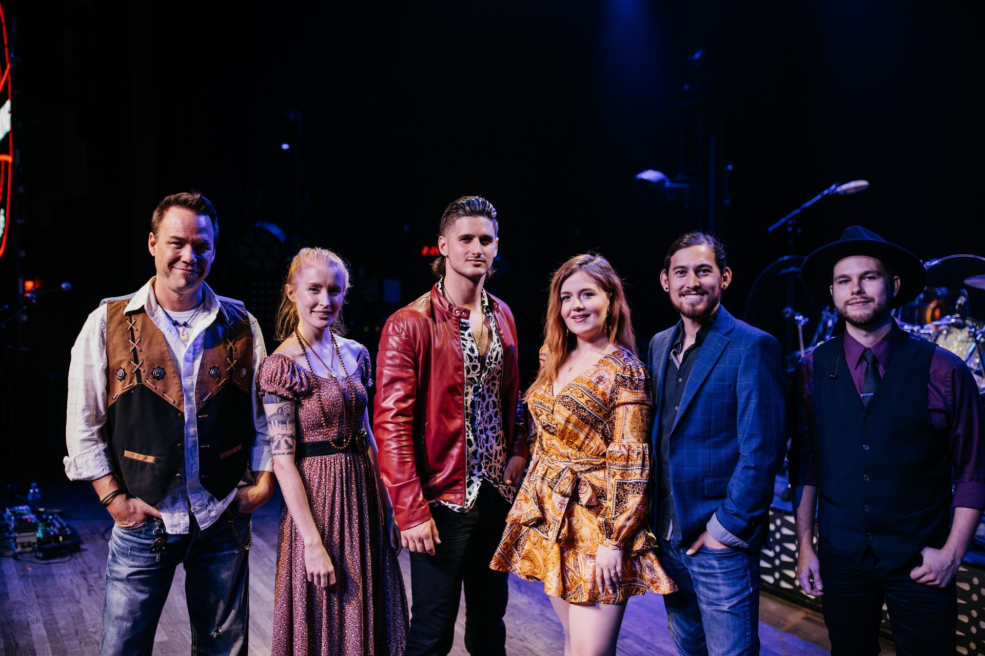 A band of six people posing together on a stage in casual and patterned outfits, with dark studio lighting.