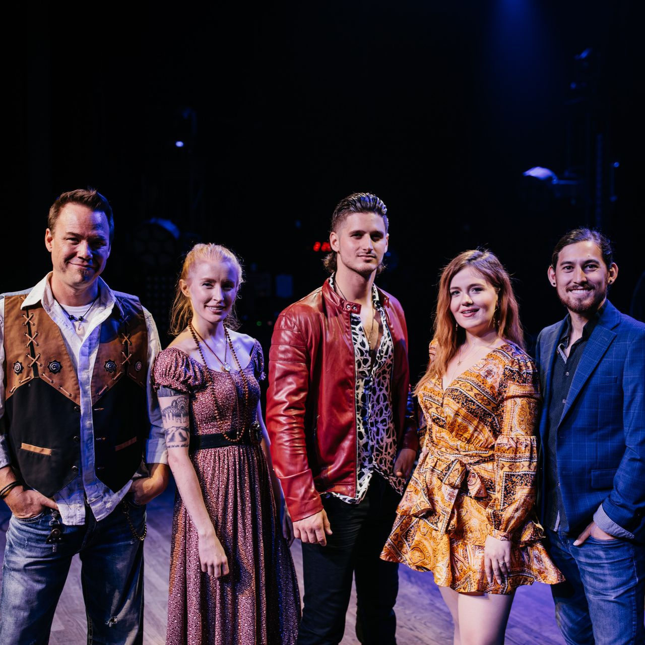 Five people stand in a line on a dimly lit stage, smiling for the camera while dressed in varied casual and formal attire.