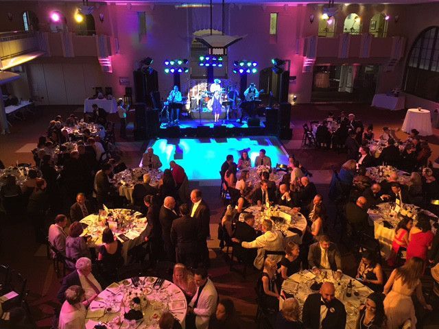 Elevated view of a ballroom with round dining tables, guests, and a brightly lit stage with a live band and dance floor.