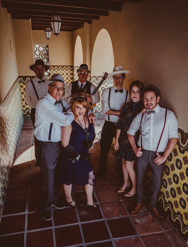 A group in 1920s attire poses in a sunlit, tiled corridor; some wear hats, suspenders, and dresses.