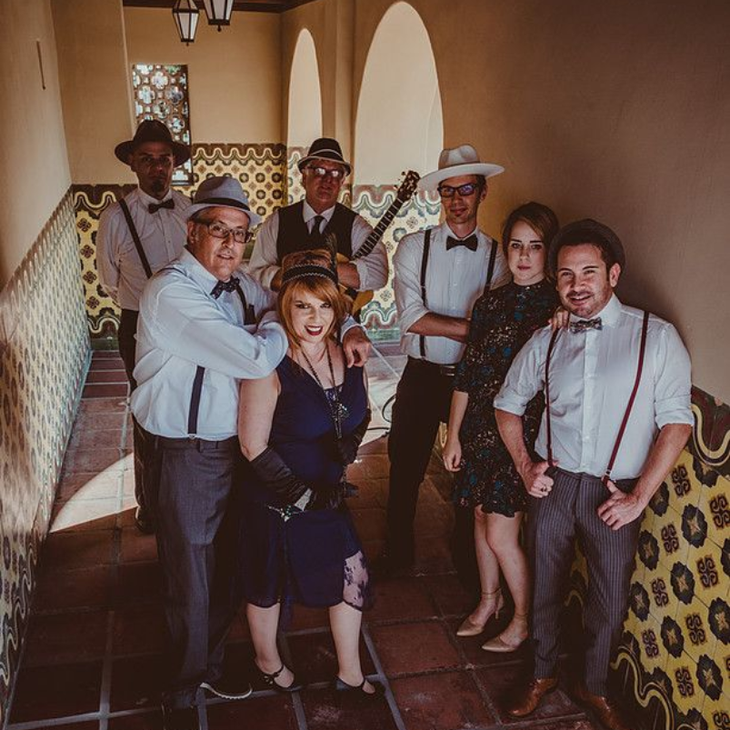 Group of eight people dressed in vintage 1920s attire posed together in an arched hallway with patterned tile walls.