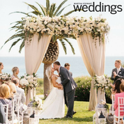 A bride and groom kiss under a draped, flower-adorned wedding arch by the ocean, with guests seated in the foreground.