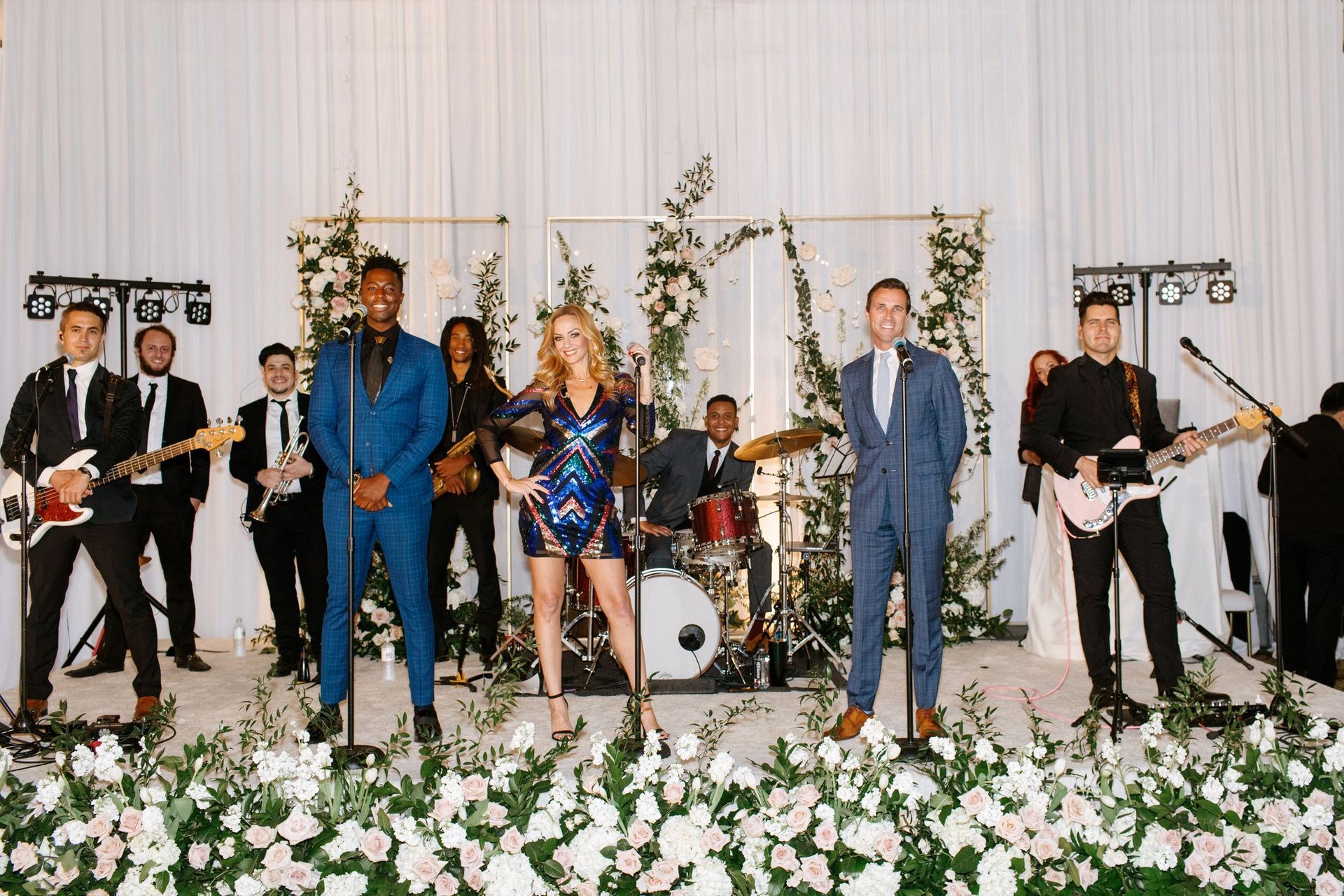 A live band performing on a stage decorated with white curtains and floral arrangements, standing before a light background.
