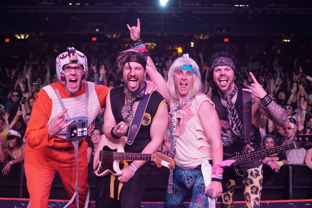 Four musicians in theatrical outfits and wigs pose on stage, smiling and making rock-and-roll hand signs to a crowd.