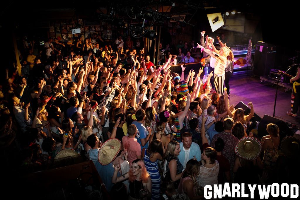 A crowded concert venue with audience members raising their hands toward performers on a dimly lit, purple-hued stage.