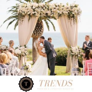 A bride and groom kiss under a floral-adorned archway at an outdoor coastal wedding ceremony.