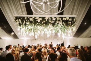 Crowd dancing in a brightly lit, white-draped tent beneath a large floral chandelier hanging from the ceiling.