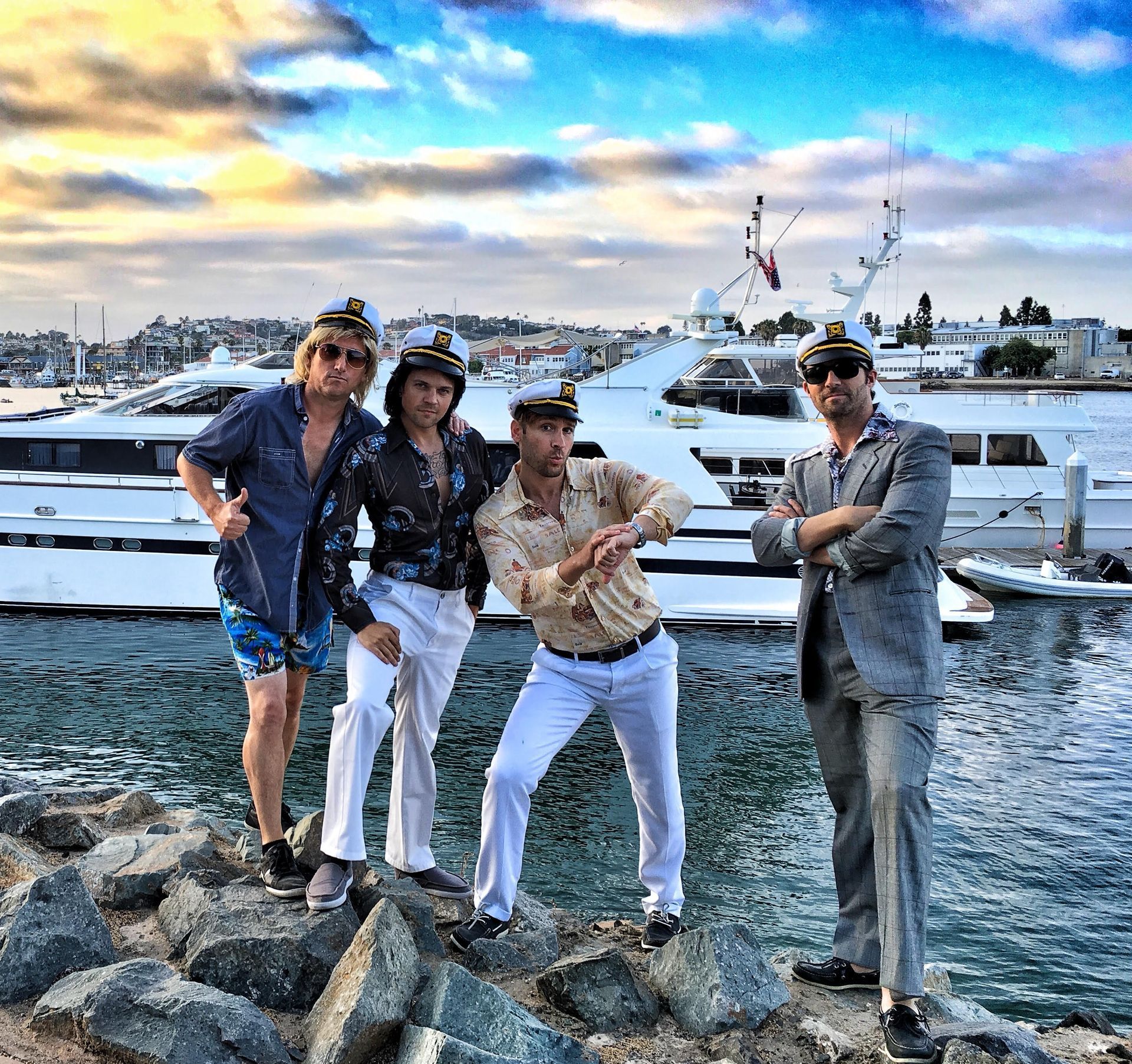Four people in captain hats pose on rocks by a marina with yachts in the background under a blue sky at sunset.