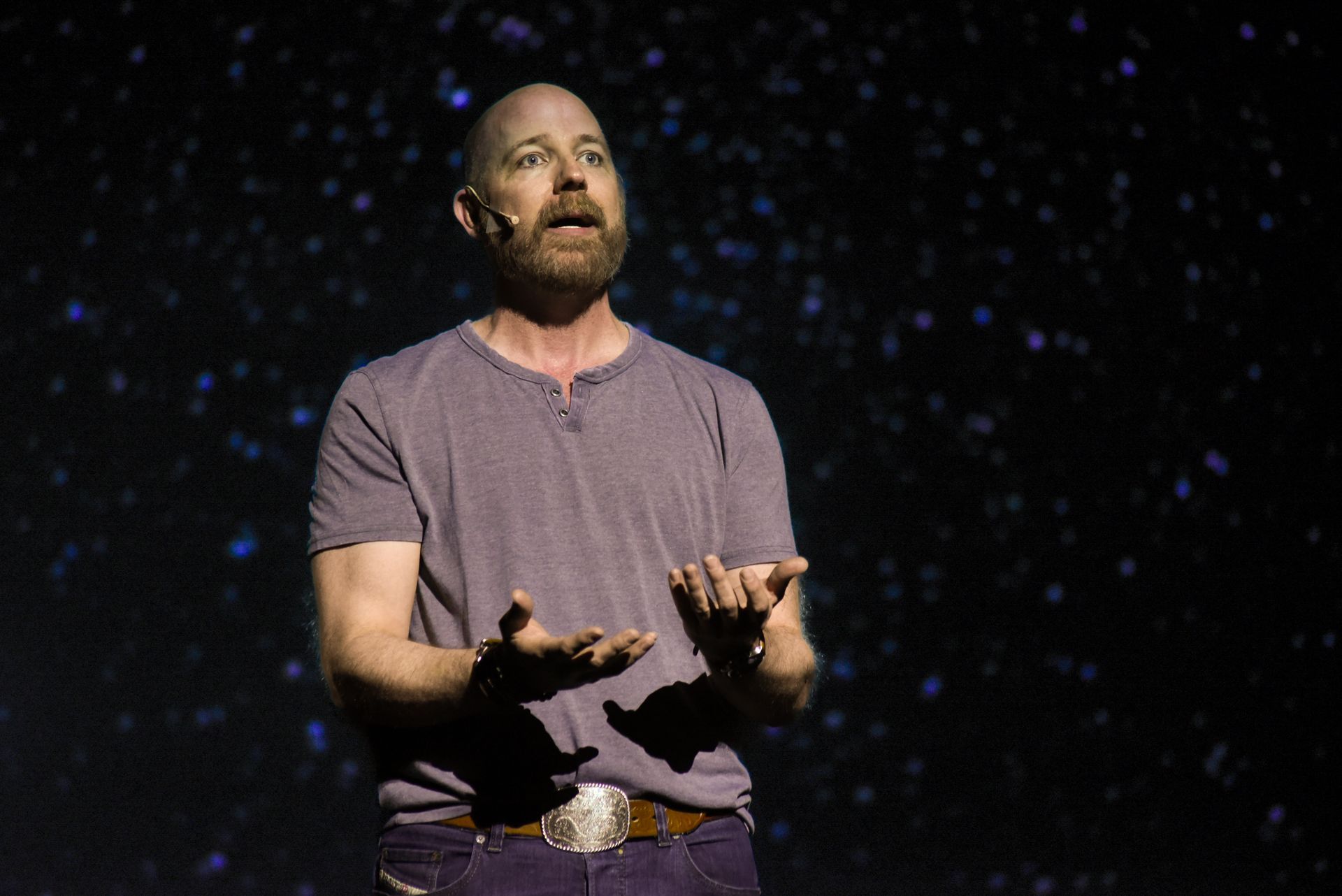 Man on stage, hands raised, speaking; purple shirt, dark background.