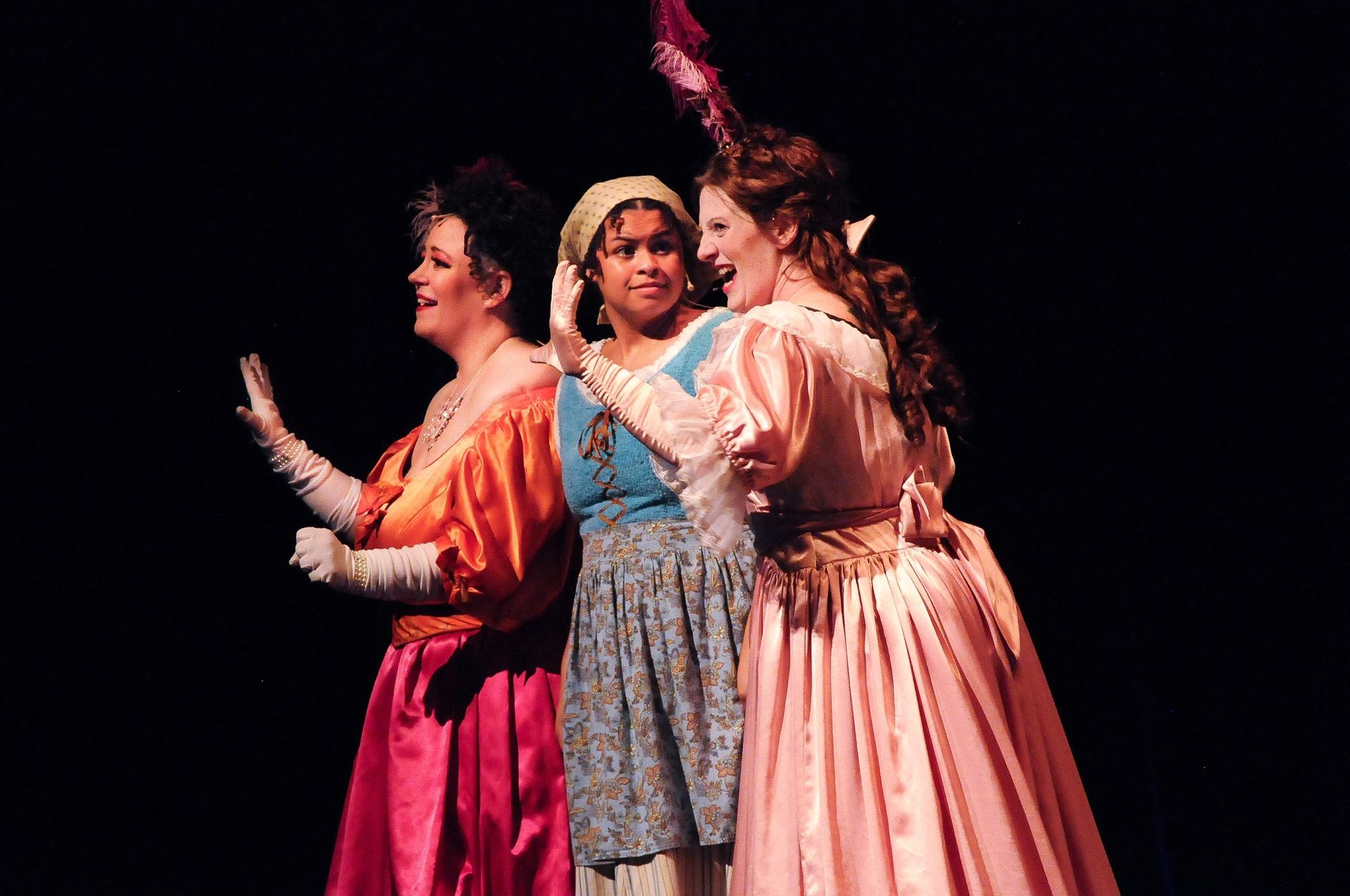 Three women in costume on stage: two in bright dresses flank a woman in blue, all smiling.