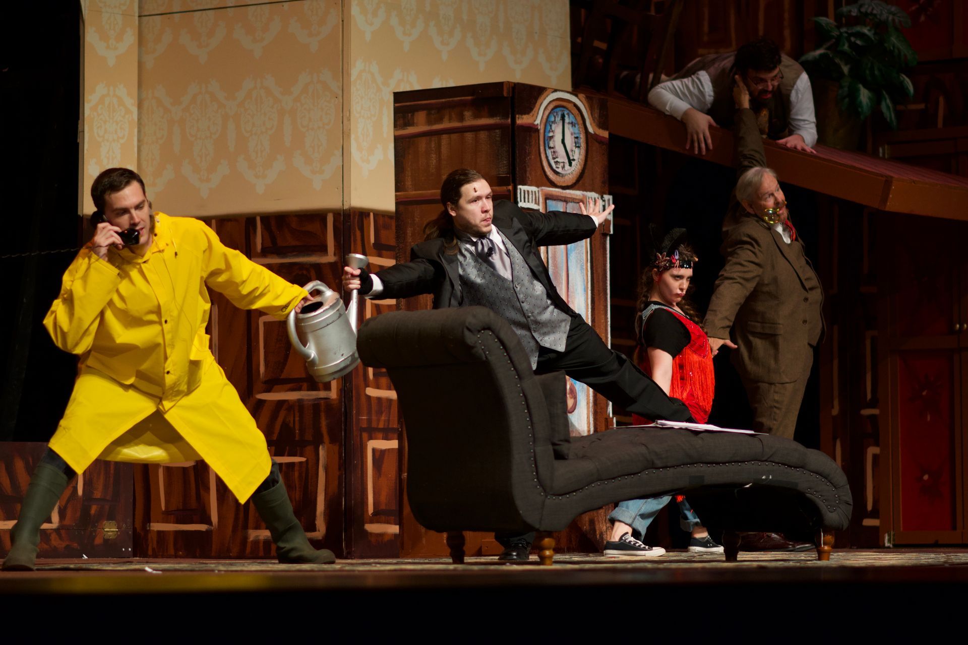 Actors on stage in a chaotic scene. A man in yellow talks on the phone, another jumps on a chaise lounge.