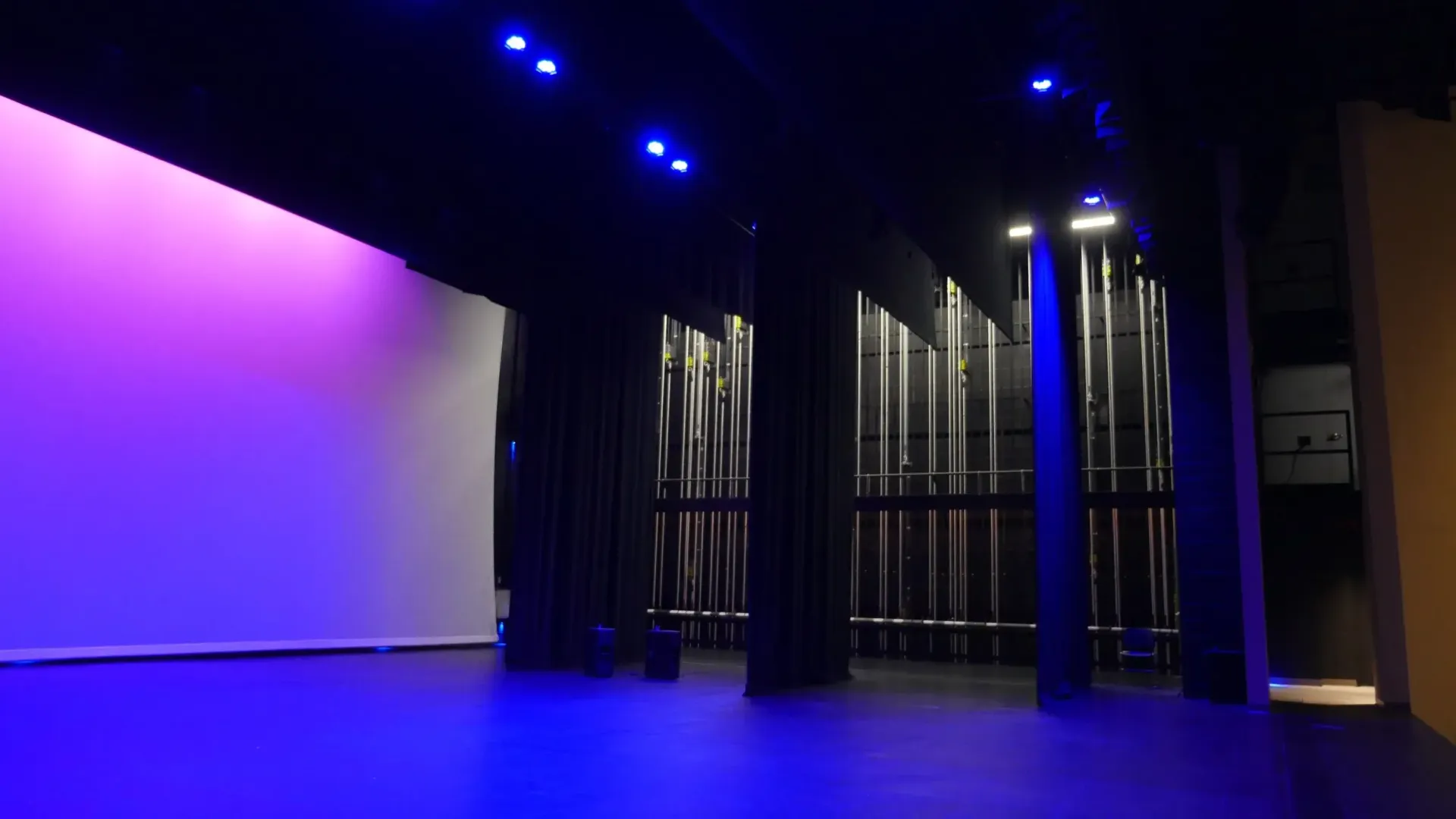 Empty theater stage with purple and blue lighting; dark curtains and stage left wall are white.