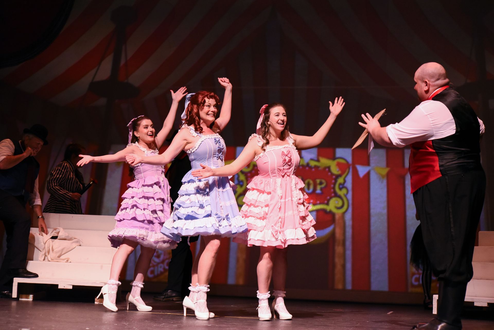 Women in colorful dresses dance on stage with a man in a circus-themed set.