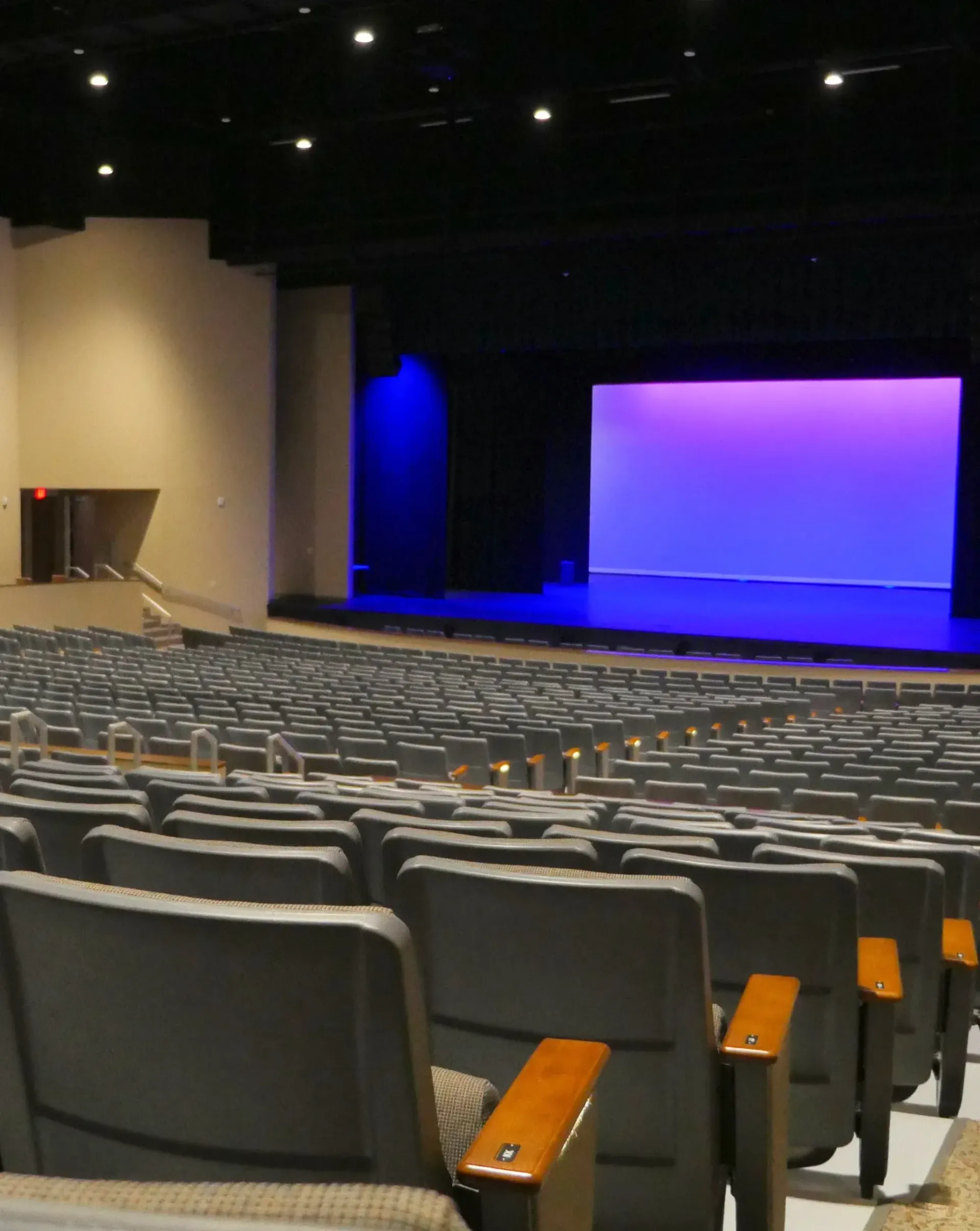 Auditorium with rows of gray seats facing a stage lit with purple and blue light.
