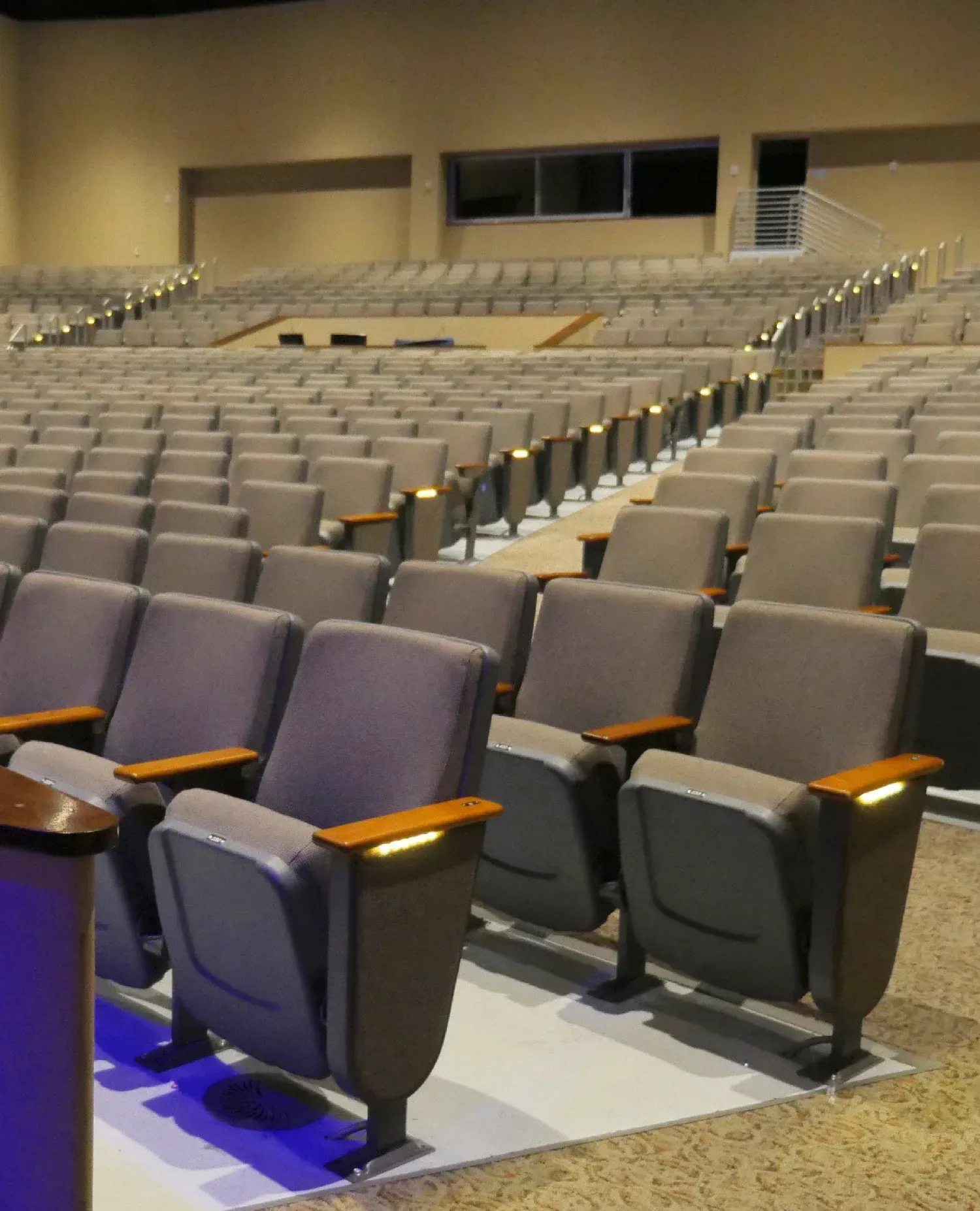 Rows of empty gray theater seats, with wooden armrests, ascending in a large auditorium.
