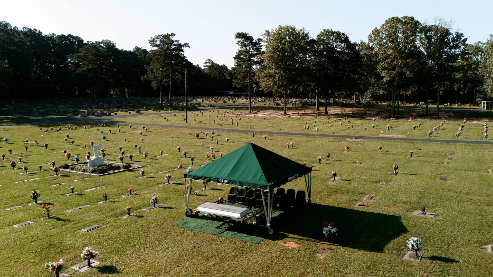 A green tent set up for a funeral service in a large, grass-covered cemetery with scattered headstones.