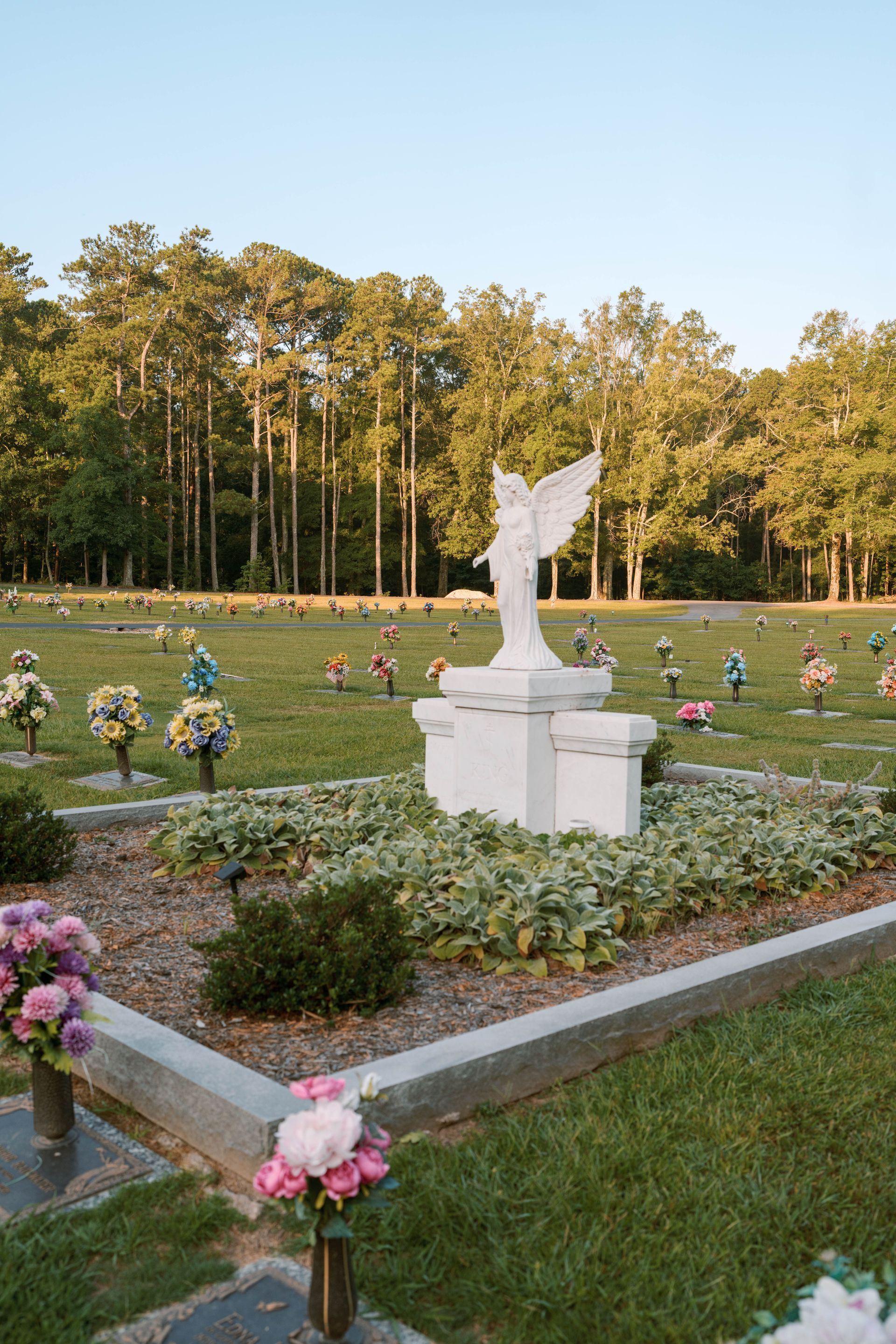 A white angel statue on a pedestal centered in a cemetery plot surrounded by flowers and trees at sunset.