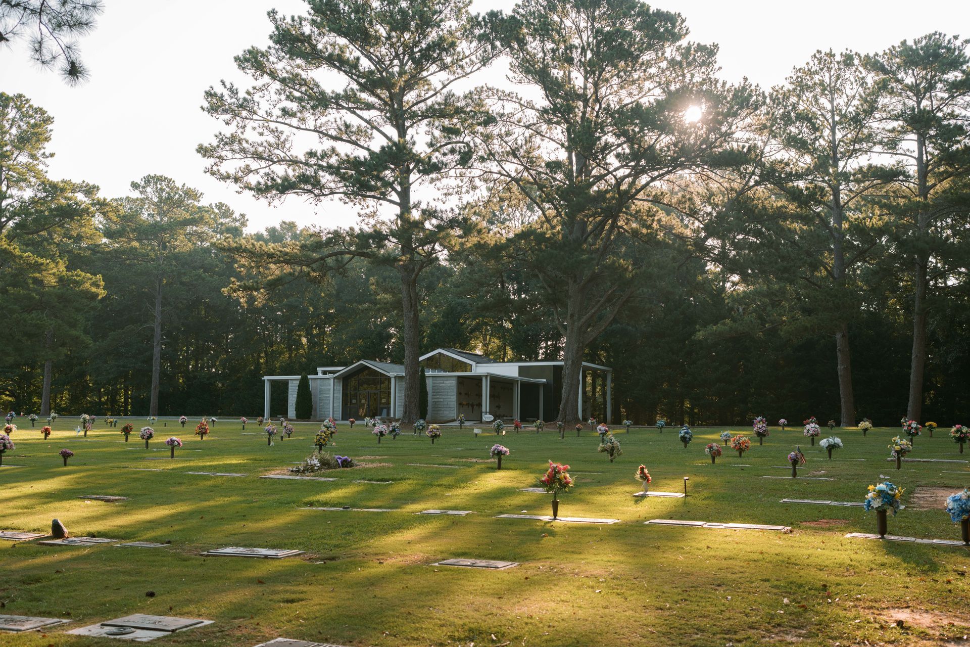 A sunlit cemetery scene with grave markers, small flower arrangements, and trees surrounding an open-air stone structure.