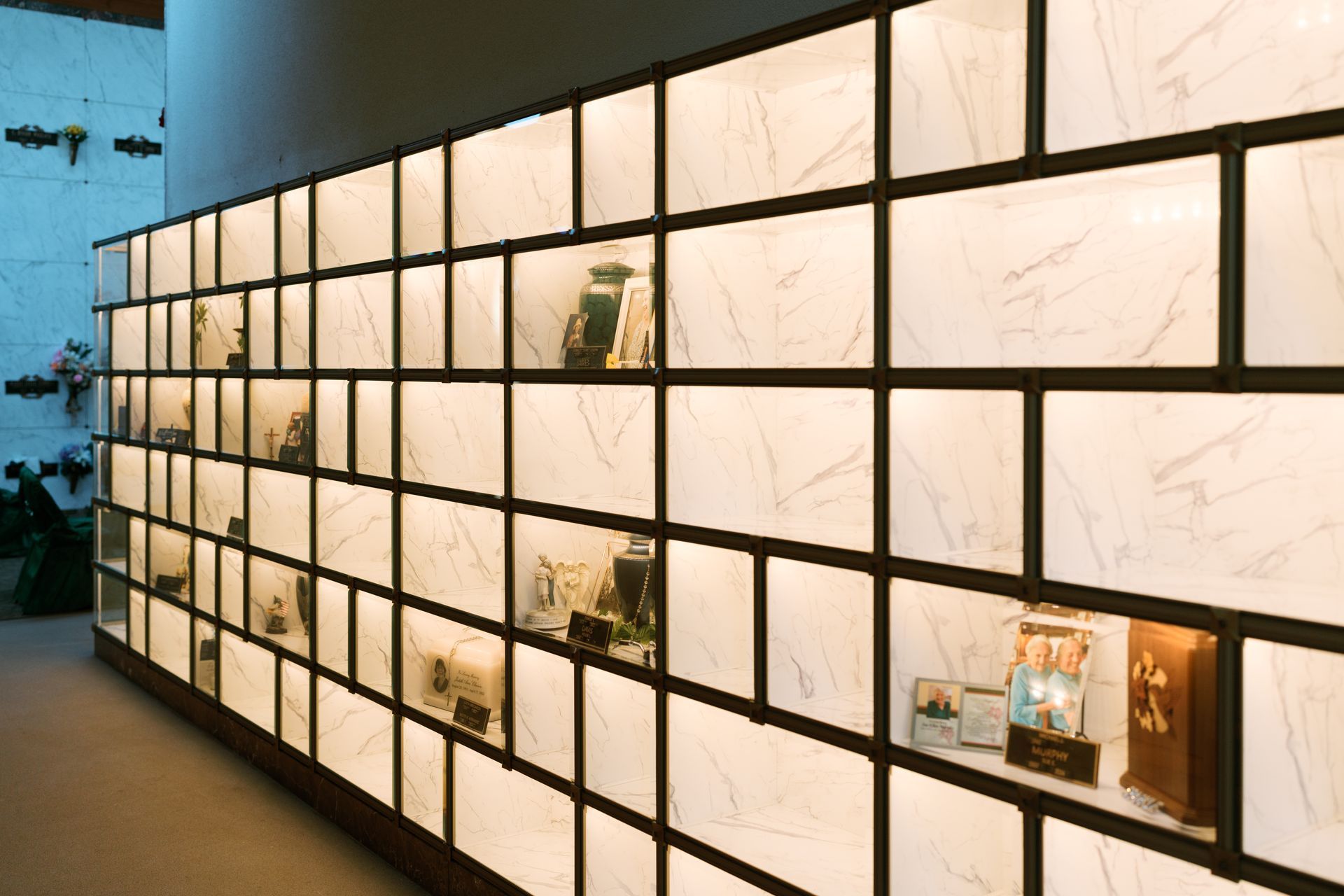 A long, illuminated indoor columbarium wall with rectangular glass-fronted niches displaying small mementos and urns.