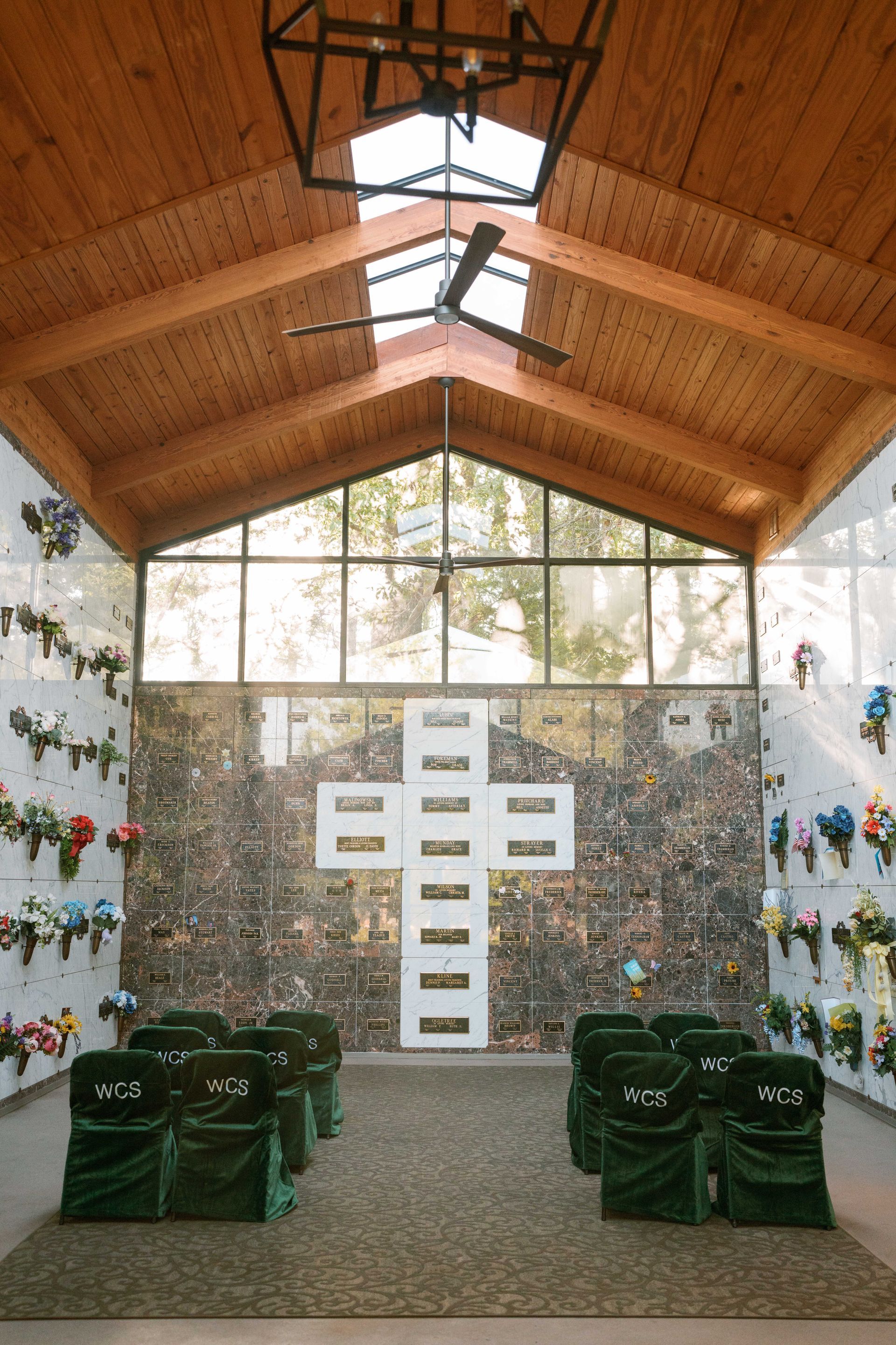 A quiet mausoleum chapel with a cross-shaped arrangement of burial niches, wooden ceiling, and rows of green-covered chairs.