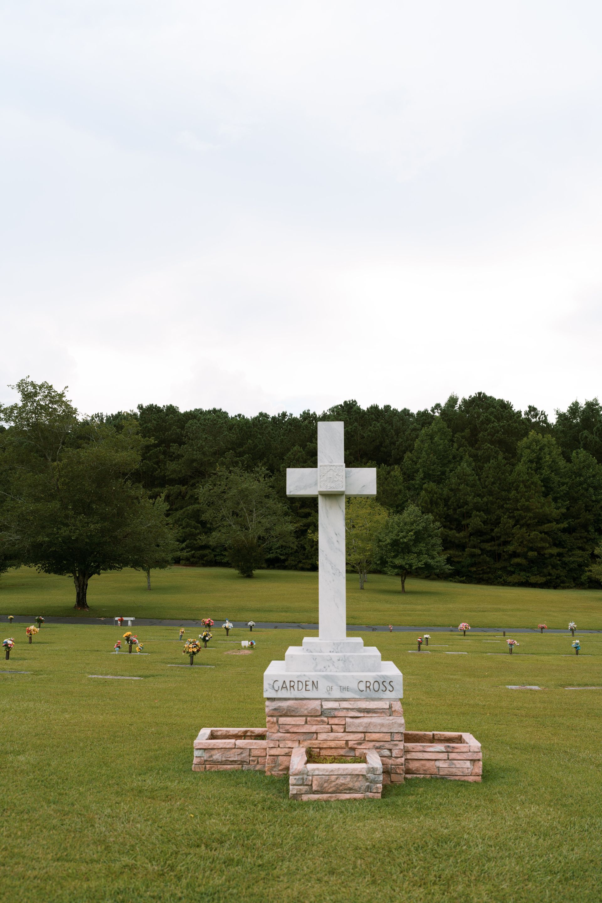 A white stone cross monument stands in a grassy cemetery, with trees in the background under a pale, overcast sky.