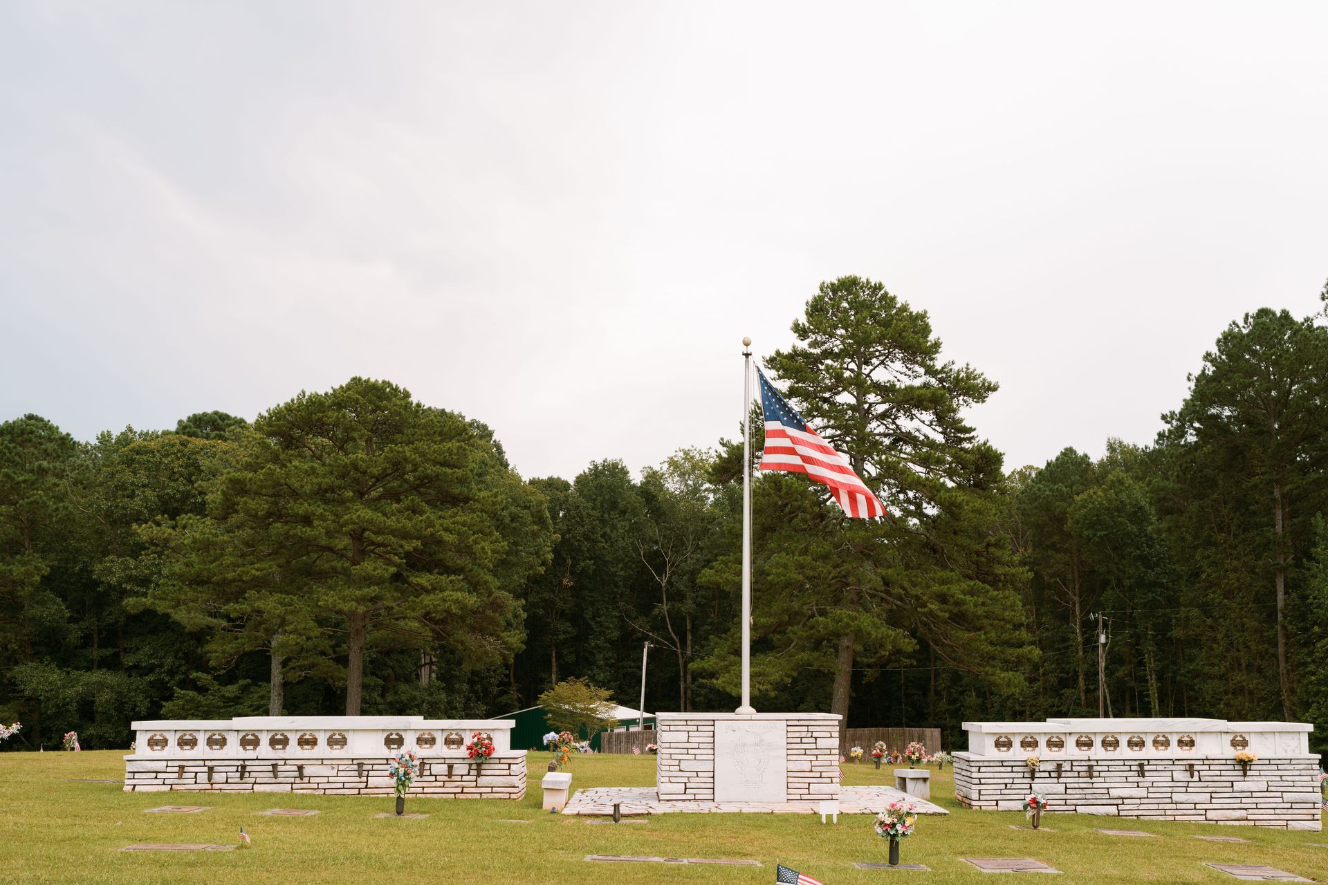 A US flag waves above a cemetery with white memorial walls, set against a background of lush green trees under a cloudy sky.