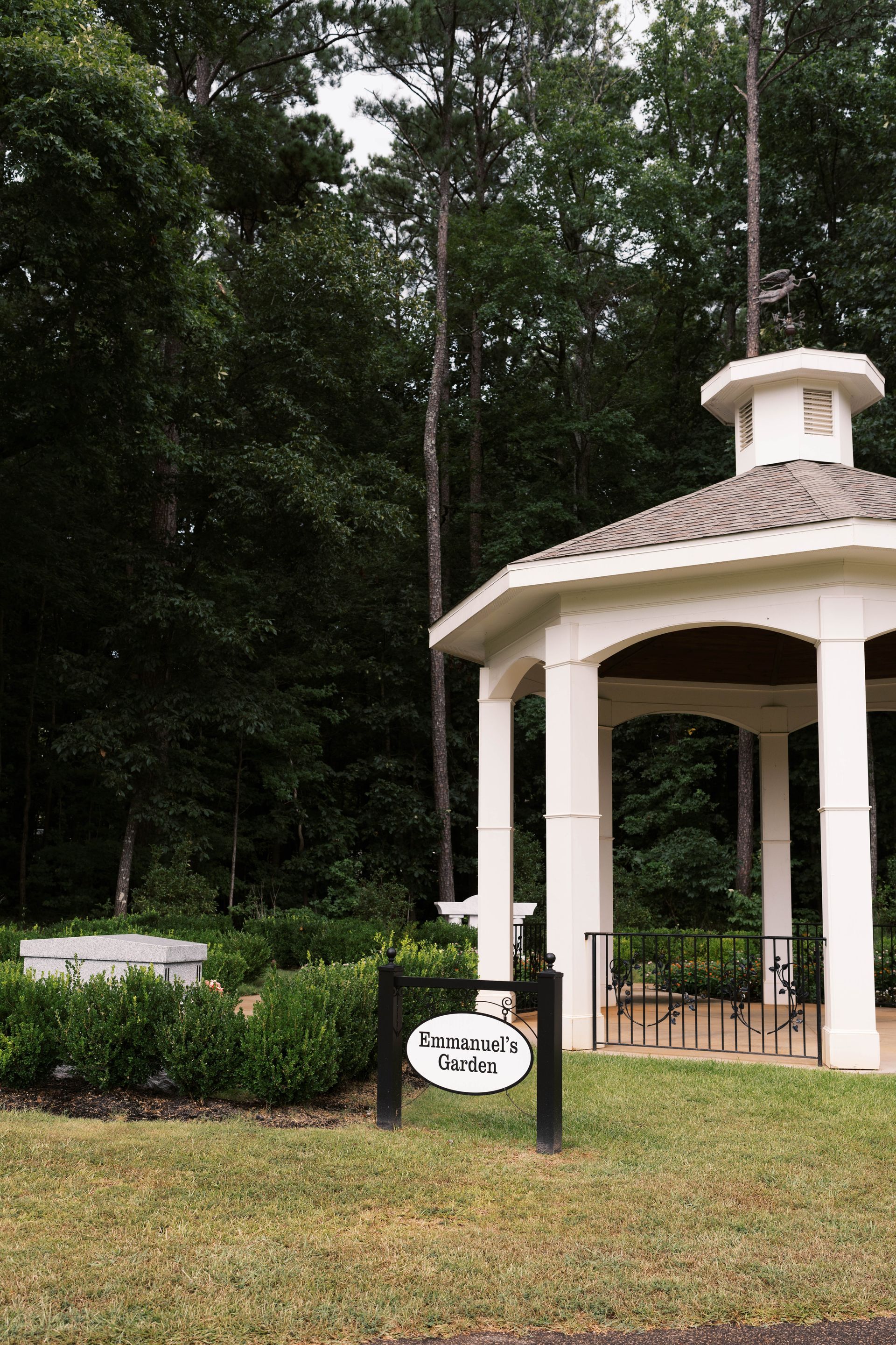A white gazebo stands in a park-like setting next to a small garden and a sign labeled