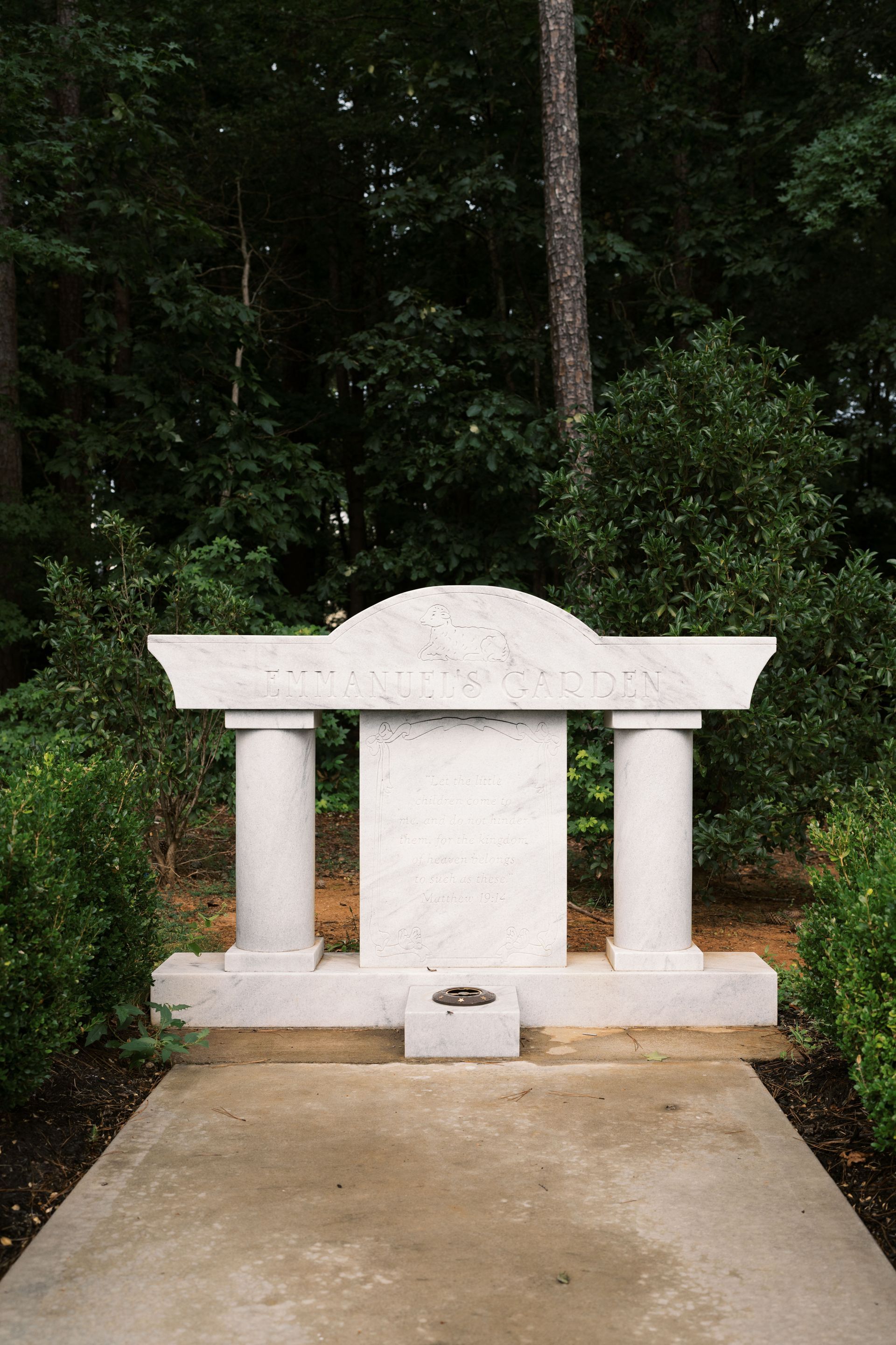 A stone monument with two columns and a central plaque sits on a concrete path in a wooded area.