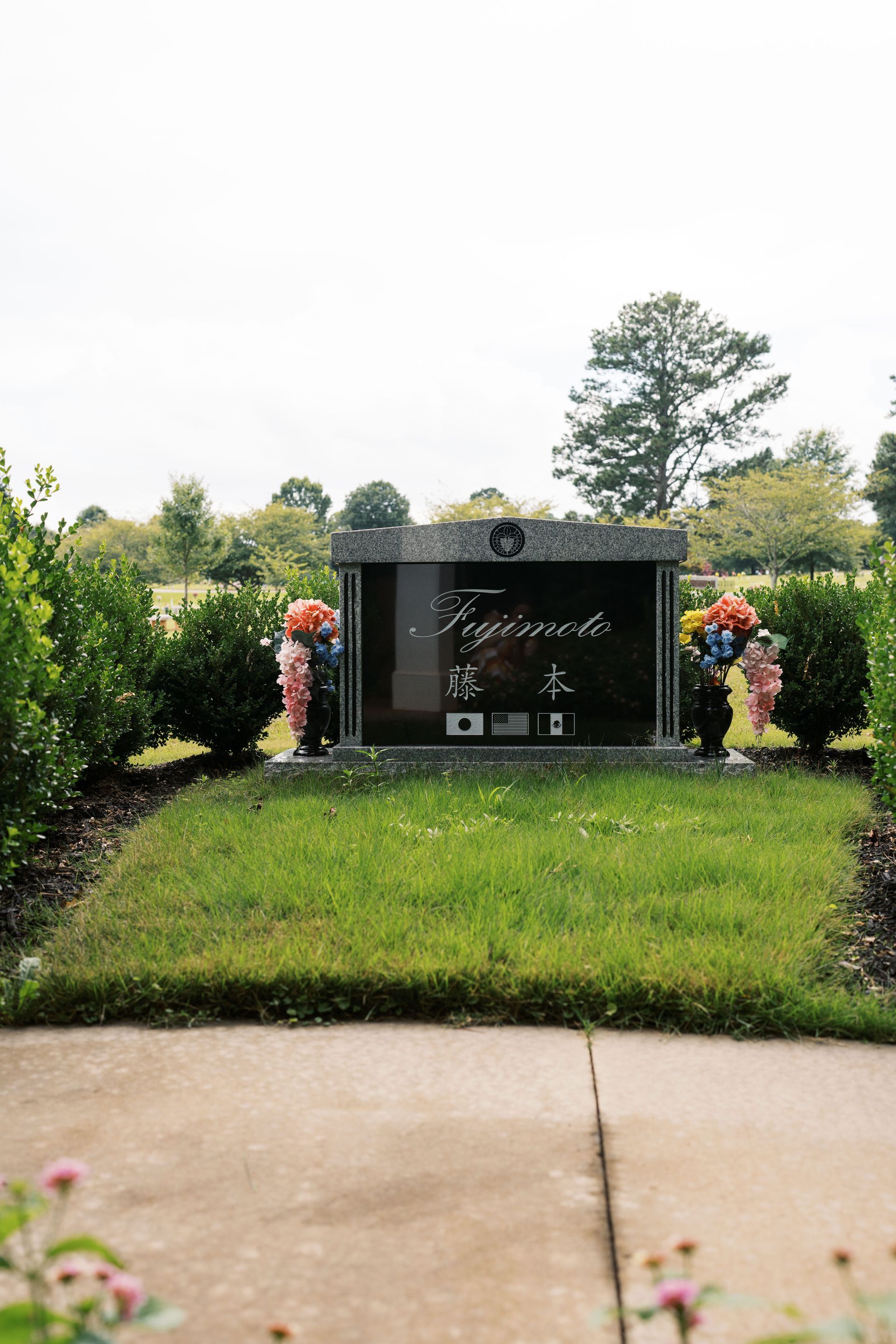 A black granite headstone with white lettering stands in a grassy cemetery plot, flanked by two small bouquets of flowers.
