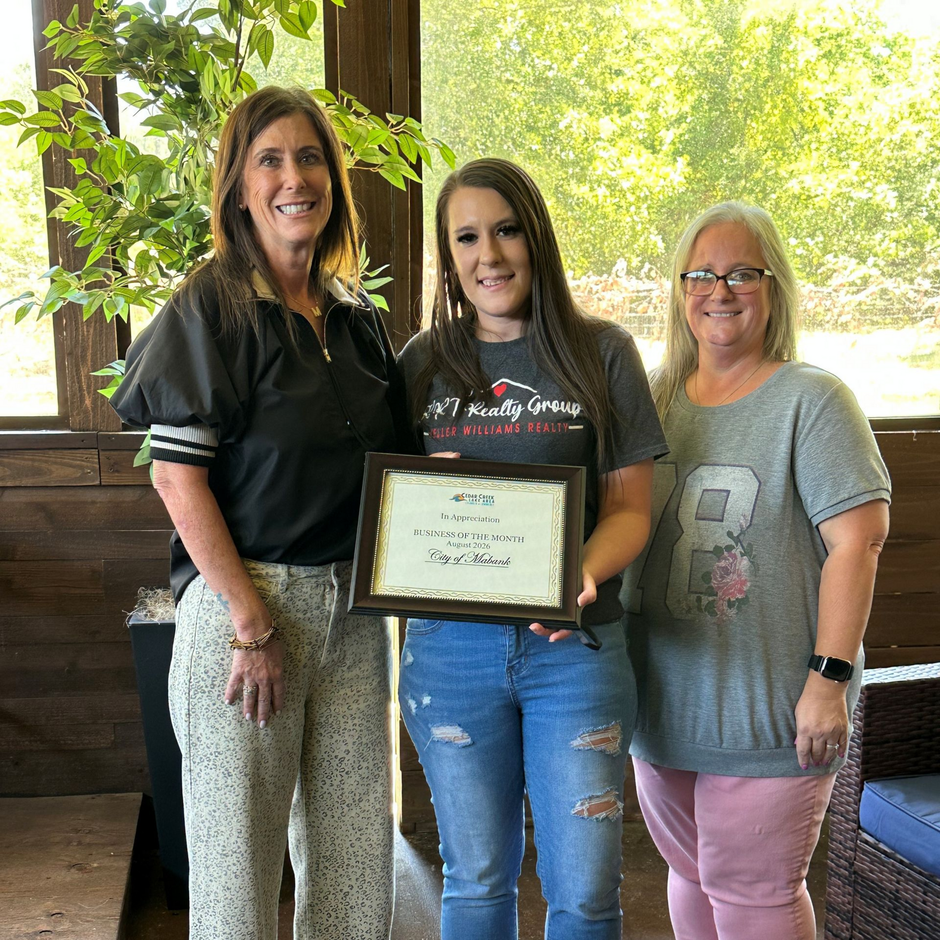 Three people smiling indoors, holding a framed certificate.
