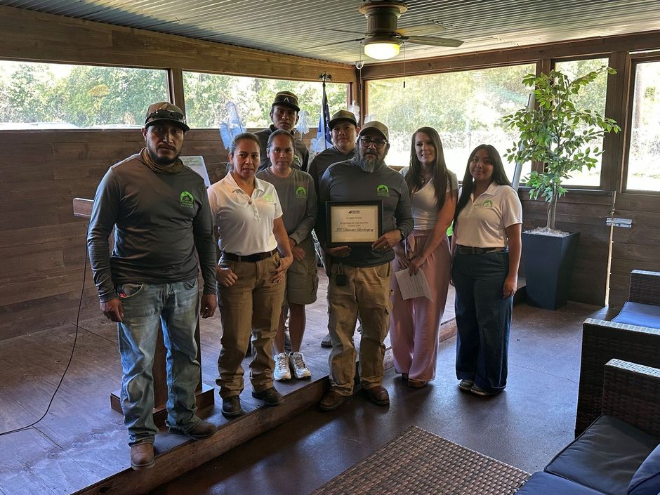 Group posing indoors with a framed certificate, standing in a rustic pavilion with trees visible outside.