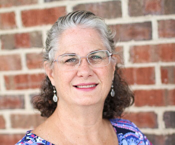 A woman wearing glasses and earrings is smiling in front of a brick wall.