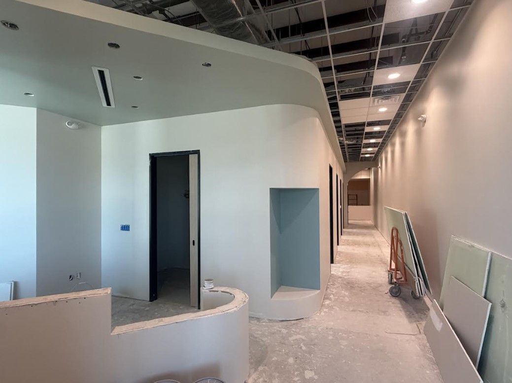 Curved hallway under construction with white walls, exposed ceiling, open doorways, and tools along the floor.
