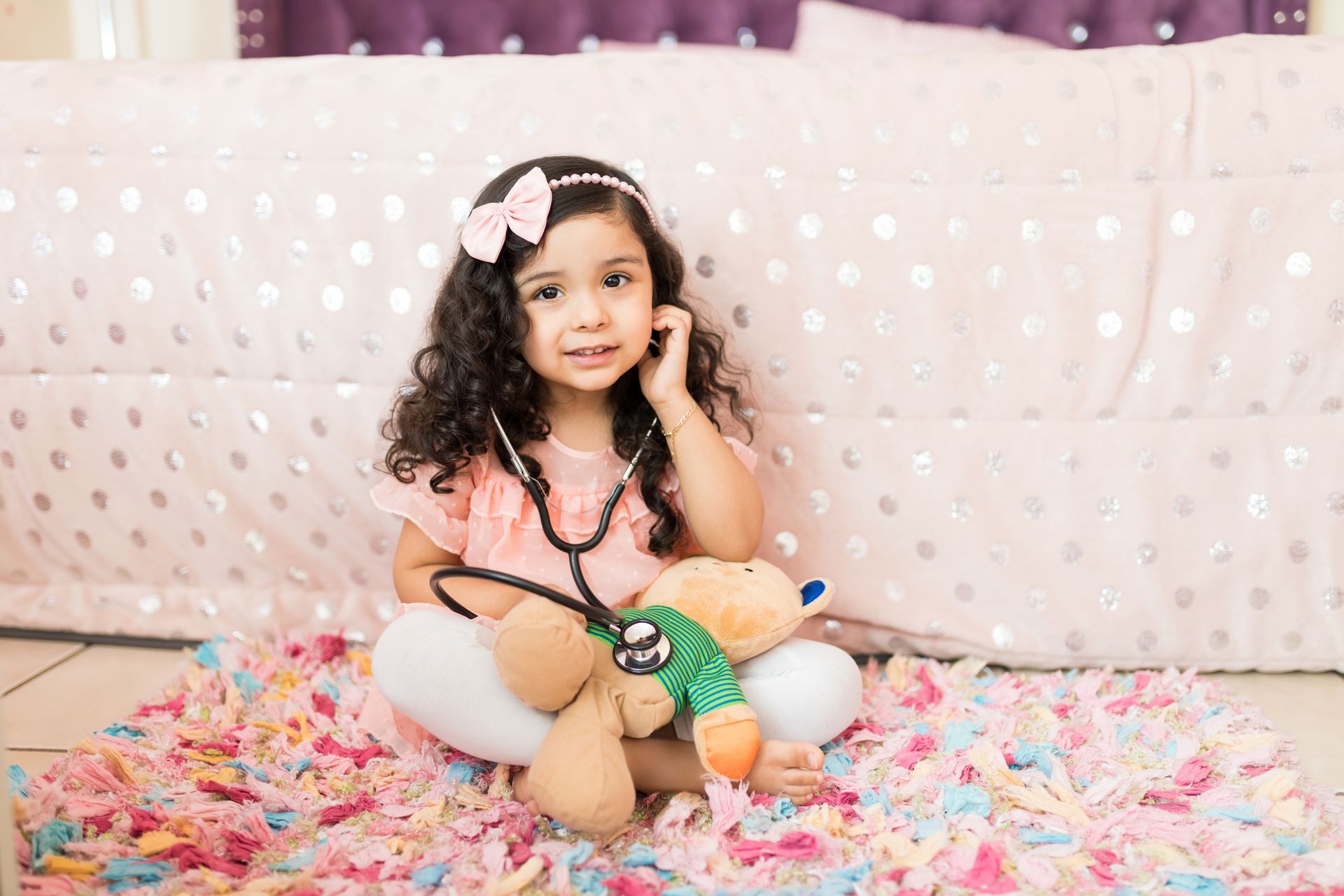 Child sitting on a flower-strewn bed, holding a teddy bear, with a pink backdrop and soft pastel decor