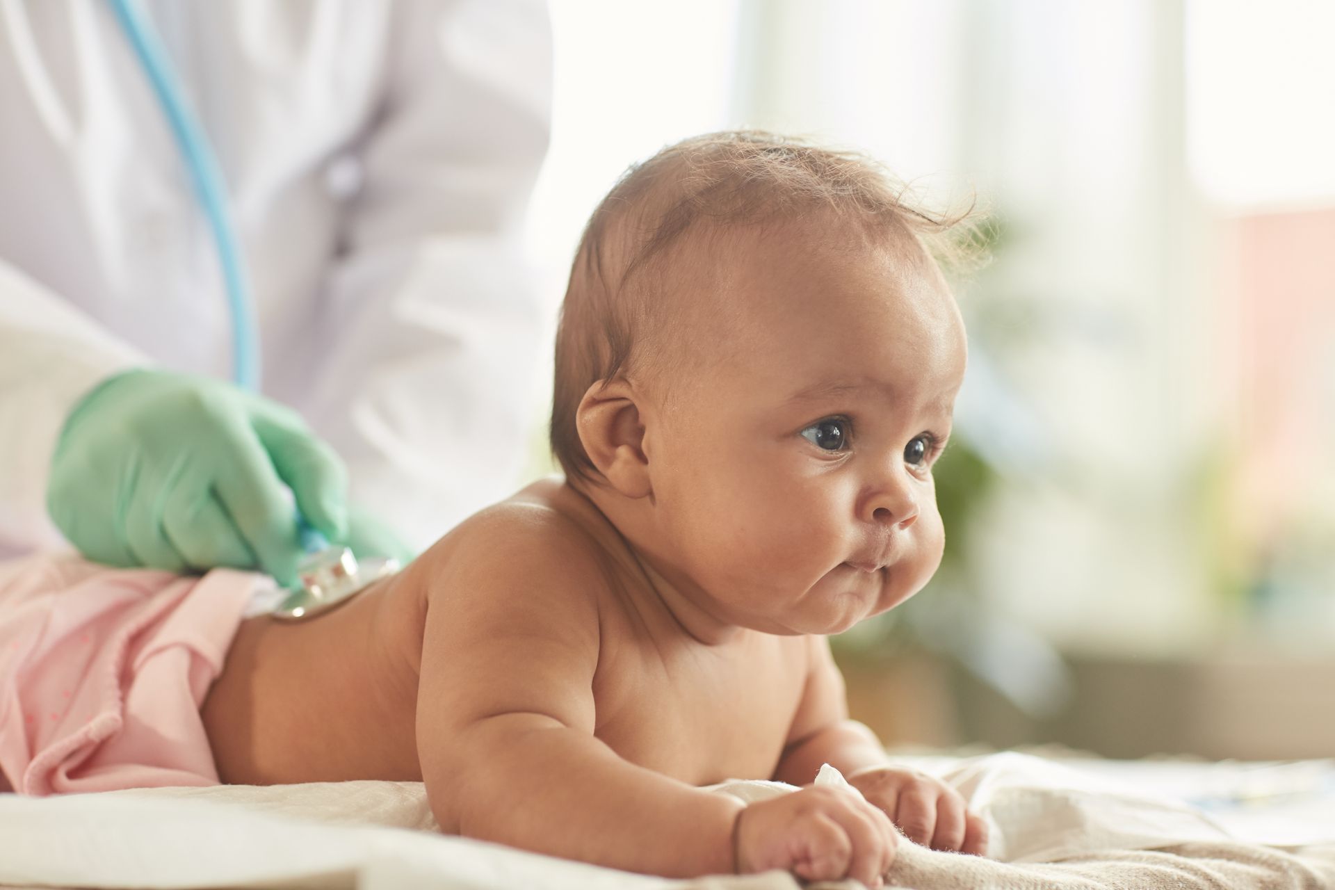 Baby lying on a medical exam table while a healthcare worker in gloves checks them with a stethoscope
