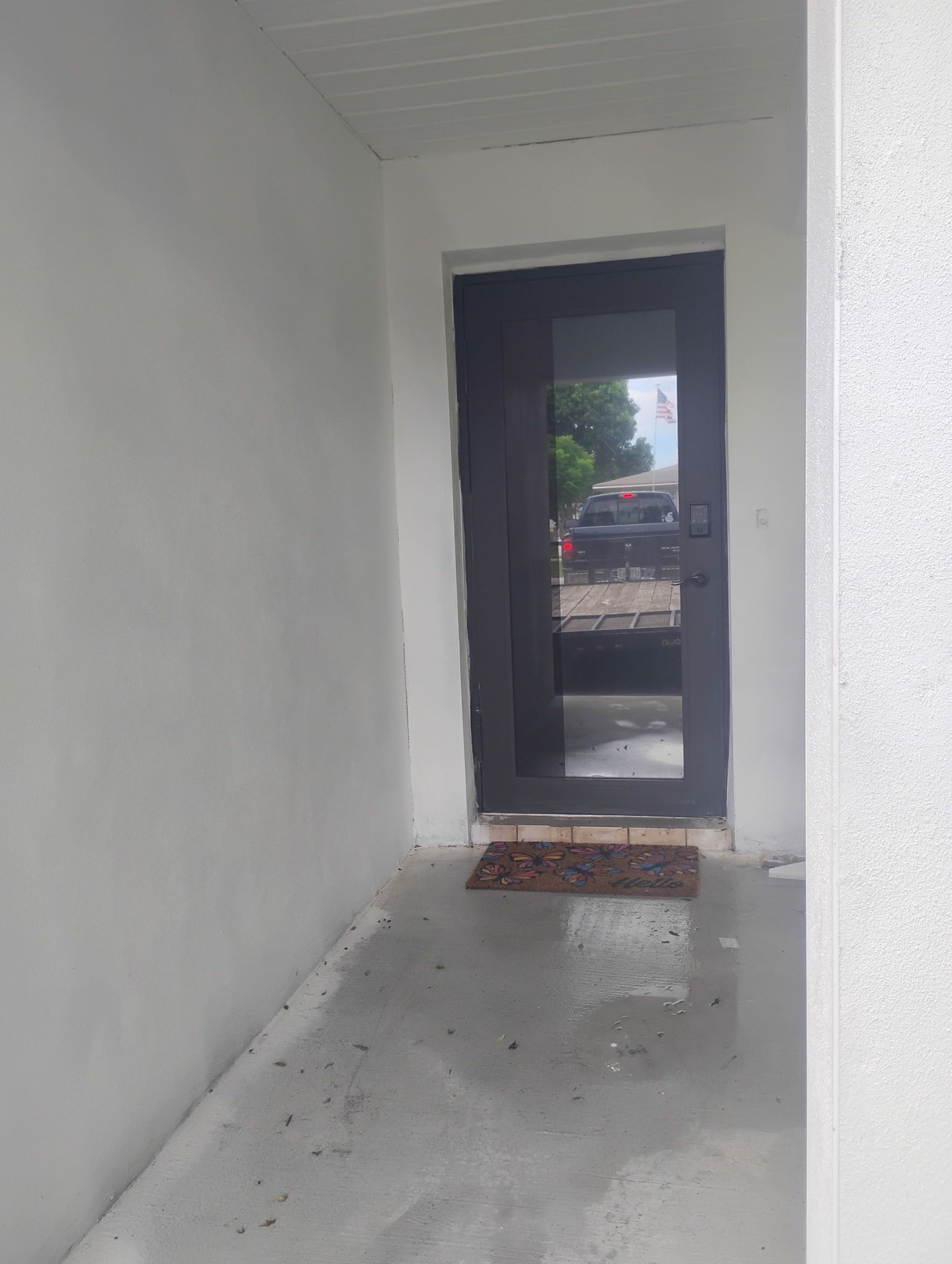 A dark door with a glass panel is set in a white exterior doorway. A welcome mat is on the wet floor.