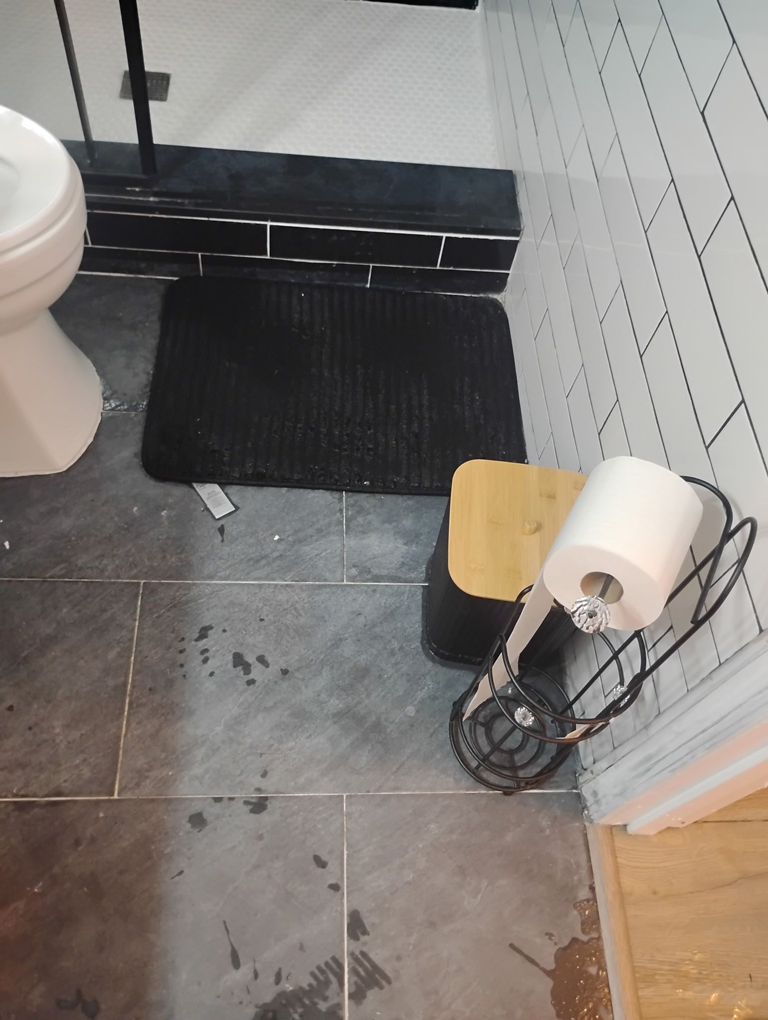 Bathroom floor with toilet, mat, small stool, and toilet paper holder. Gray tile floor with white patterned wall.