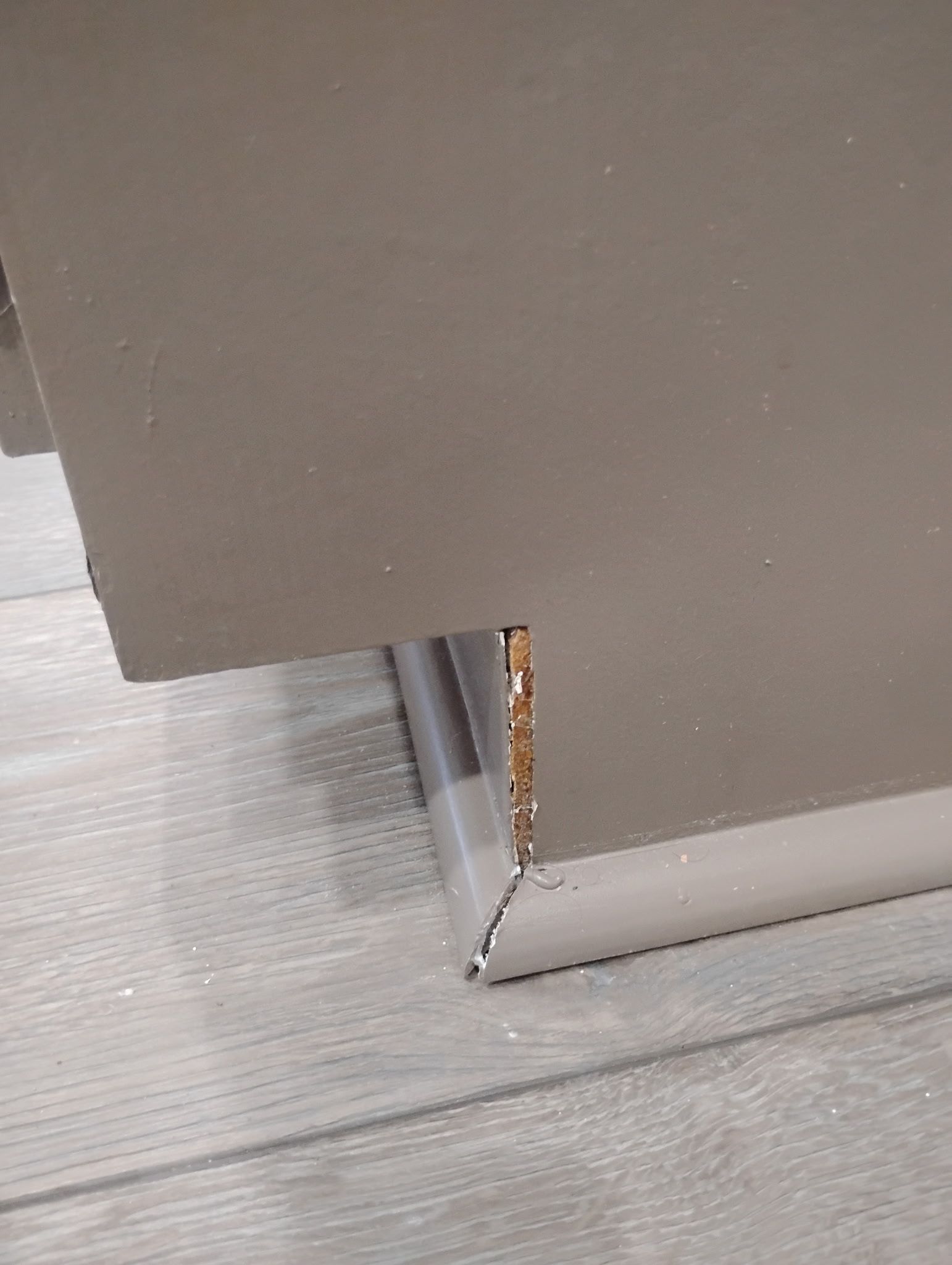 Gray cabinet edge meets baseboard, revealing raw wood. Silver baseboard on a light gray floor.