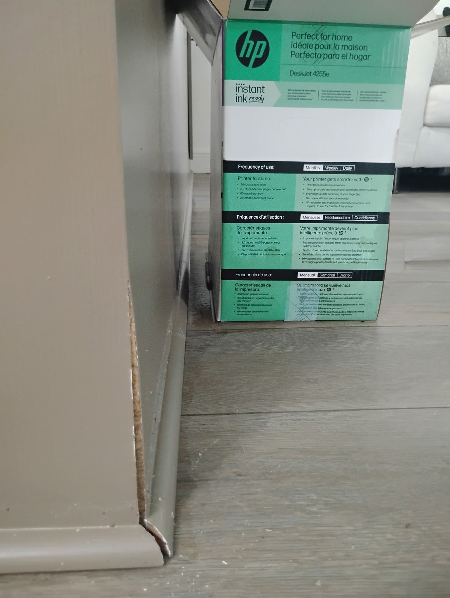 An HP printer box sits against a light-colored cabinet on a wood floor.