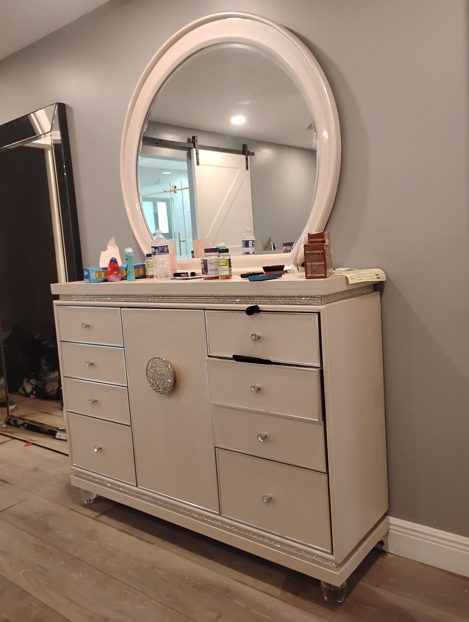 White dresser with oval mirror, set against a gray wall. Vanity with several drawers and a center decorative element.