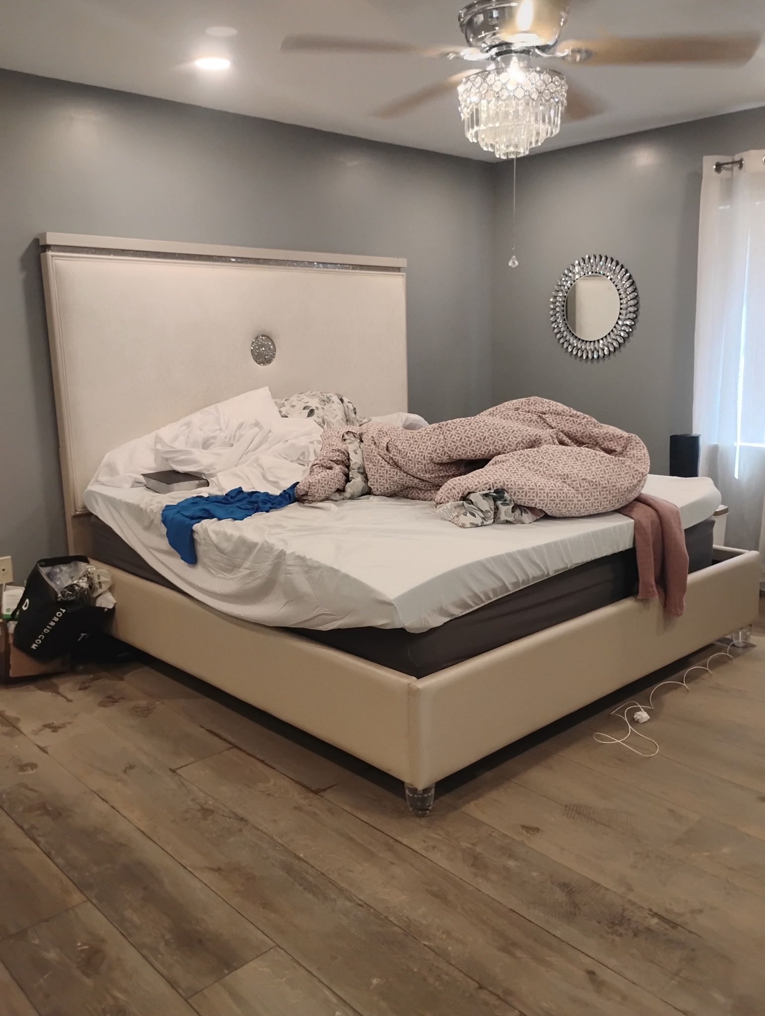 A disheveled bed with a tall, light-colored headboard in a bedroom with wooden floors and gray walls.