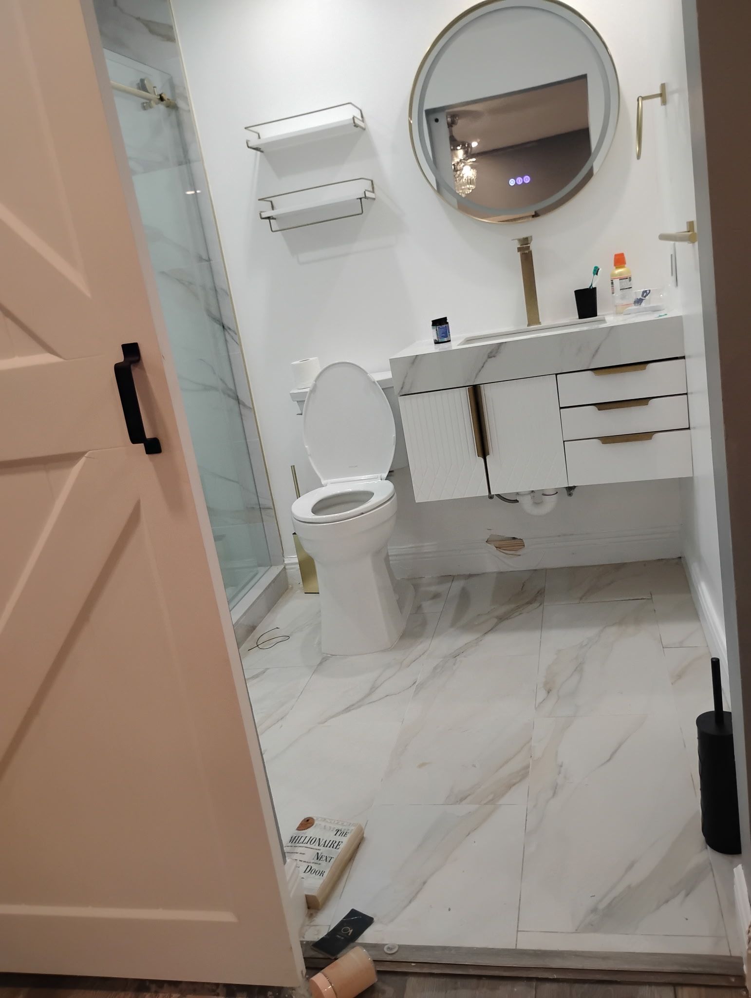 White bathroom with marble-look tile, floating vanity, round mirror, and a partially open white barn door.