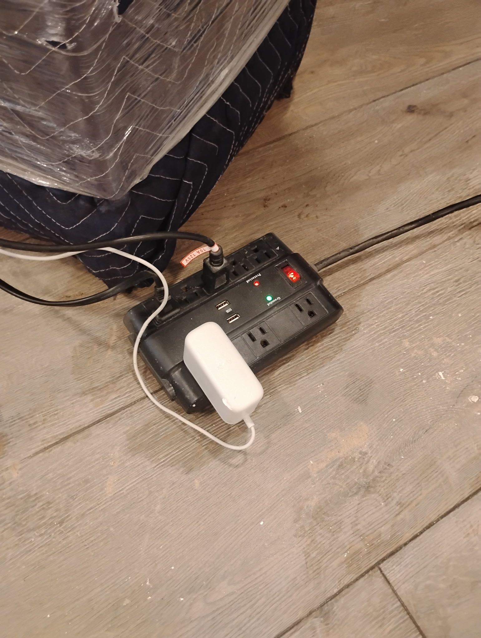 Black power strip on wood floor, with devices plugged in. A wrapped item is in background.